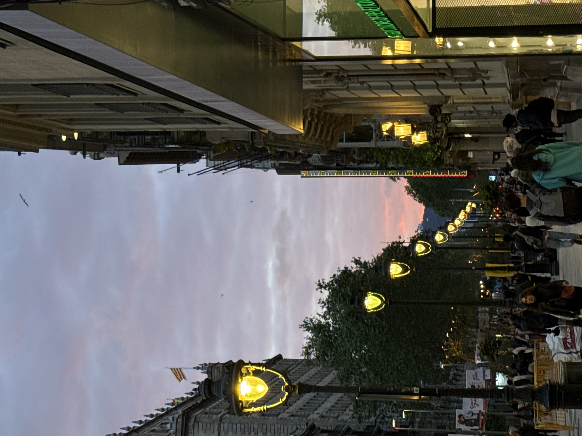 This image shows a street in Barcelona, Spain at twilight. The street is lined with ornate street lamps which are illuminated. There are people walking on the sidewalk. In the background, the sky has pink and orange hues. The architecture is grand, with buildings lining the street. A Spanish flag is visible atop one of the buildings.