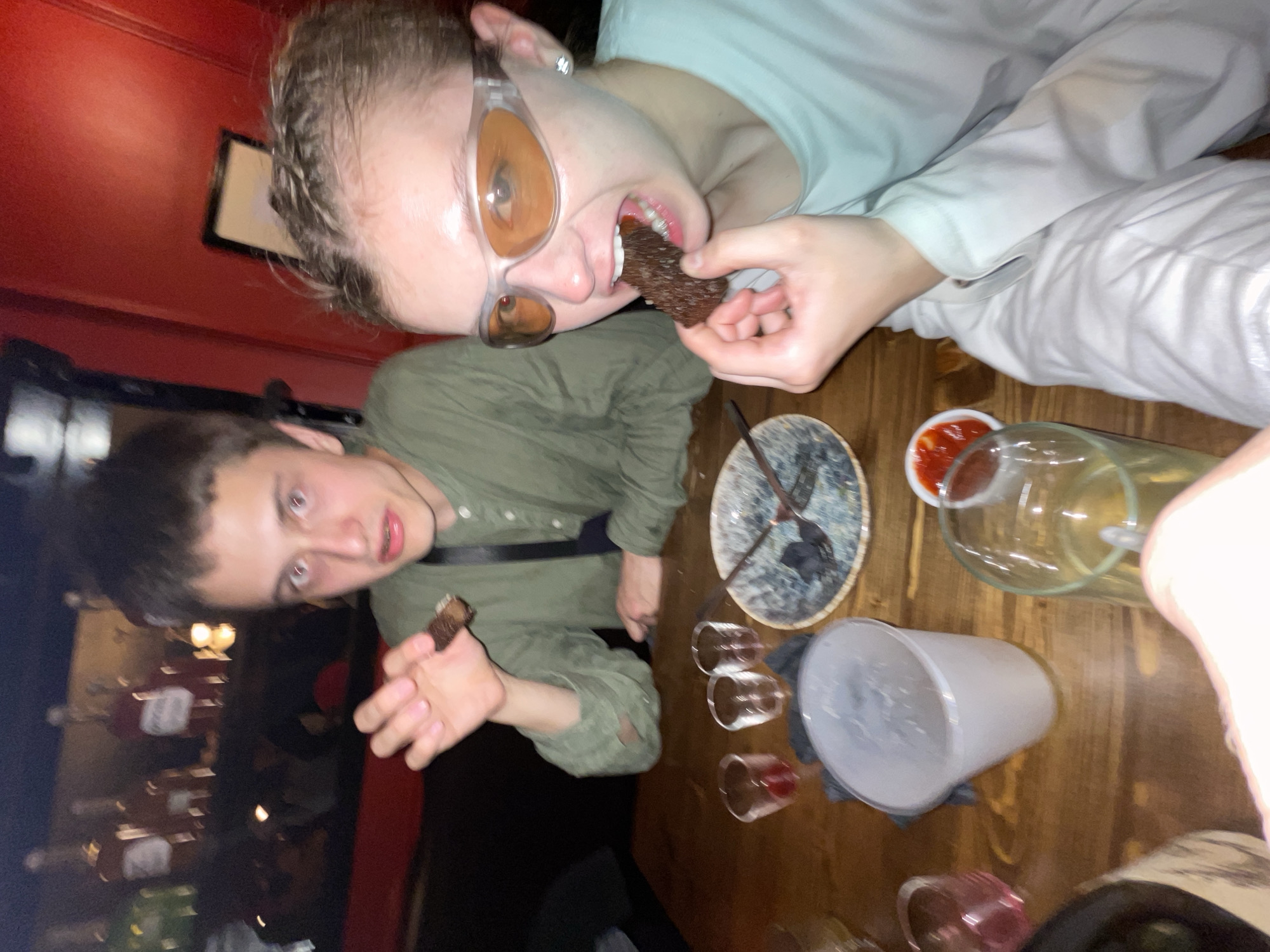 Two individuals are at a table in what appears to be a dimly lit restaurant or bar. One person is wearing translucent framed sunglasses and eating a piece of dark food. The other person is wearing a green button-down shirt and holding a similar piece of food. The table is wooden and has a plate, several small glasses, a larger plastic cup, a pitcher of liquid, and a small dish with red sauce. The background shows blurred details of the restaurant, including seating and lighting. The location is not specified as being in Moscow, Russia.