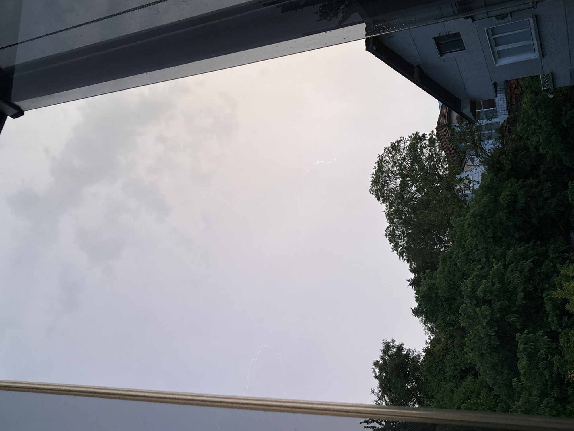 The image depicts a cloudy sky with visible lightning. A building and trees are visible on the right side of the frame. The view is framed by part of a building structure, suggesting the photo was taken from indoors, looking out through a window or opening. Given the presence of buildings and greenery, it's likely an urban or suburban area. Without further information, it's impossible to confirm the exact location in Varna, Bulgaria.
