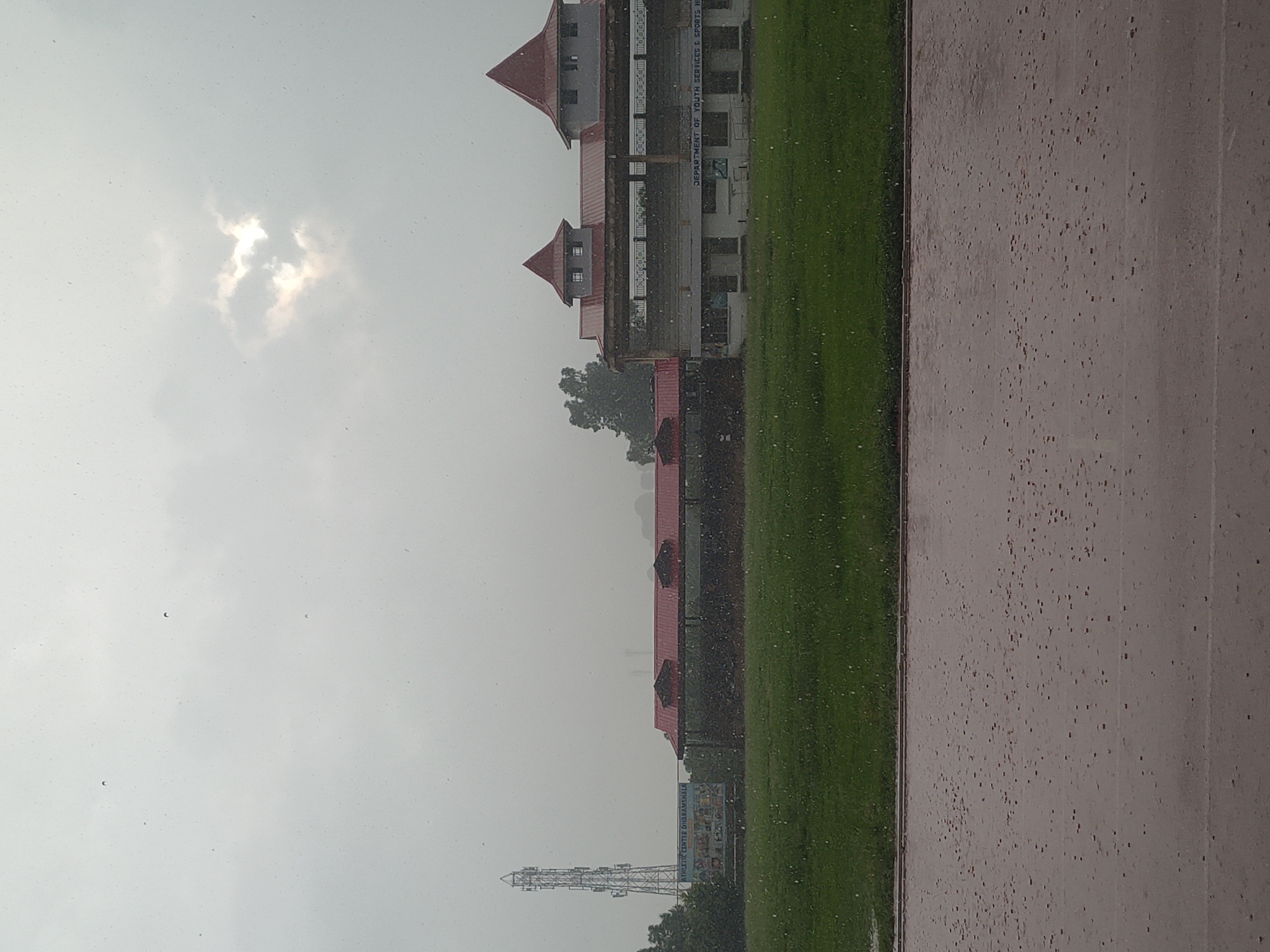 The image depicts the Himachal Pradesh Cricket Association Stadium in Dharamshala, India, during what appears to be a rainy day. The sky is overcast with a patch of sunlight breaking through clouds, casting a subtle glow. Raindrops are visible throughout the frame, particularly on the foreground track which is wet and reflects the light. A green field, likely the cricket pitch, is situated behind the track.
In the middle ground, a building with red-tiled roofs and grey walls stands prominently. The building features architectural elements like turrets and balconies. Text on the building reads "DEPARTMENT OF YOUTH SERVICES & SPORTS HP". To the left of this building, a tall telecommunications tower is visible, along with a signboard that displays "ATHLETIC CENTER DHARAMSHALA" and some images. Trees are present on the left side of the image. The overall atmosphere suggests a damp and slightly hazy environment due to the rainfall.
