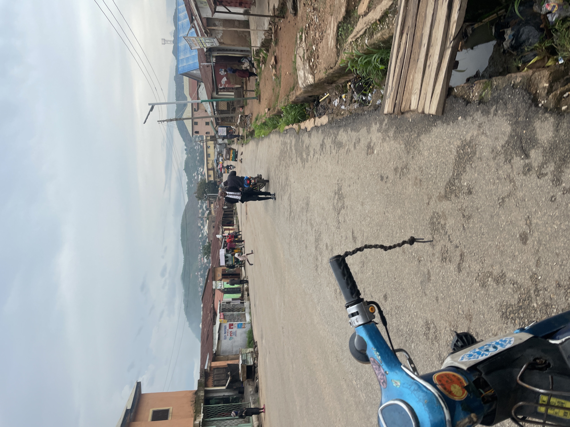 The image depicts a street scene in Jos, Nigeria, likely during daylight hours with overcast skies. The perspective is from a person on a blue scooter, whose handlebars and front portion are visible in the foreground. The street is paved and appears to be a local thoroughfare lined with buildings on both sides.
On the left side of the street, there are several buildings, some of which appear to be shops or residential structures with corrugated metal roofs and some with concrete facades. One building has an orange-toned exterior. On the right side of the street, there are also buildings with similar roofing materials. A drainage ditch runs along the side of the road on the right, containing some debris and water. A makeshift wooden bridge crosses over the ditch.
Several people are visible in the scene. Two men in suits are walking on the road, one of whom appears to be passing another individual on a motorcycle. Further down the street, other individuals are seen walking or standing. In the middle ground, a child is running across the road. In the background, rolling hills covered with scattered settlements can be seen under the cloudy sky. Electrical wires are strung across poles, indicating the presence of infrastructure. The overall impression is a busy, everyday scene in an urban or semi-urban environment.