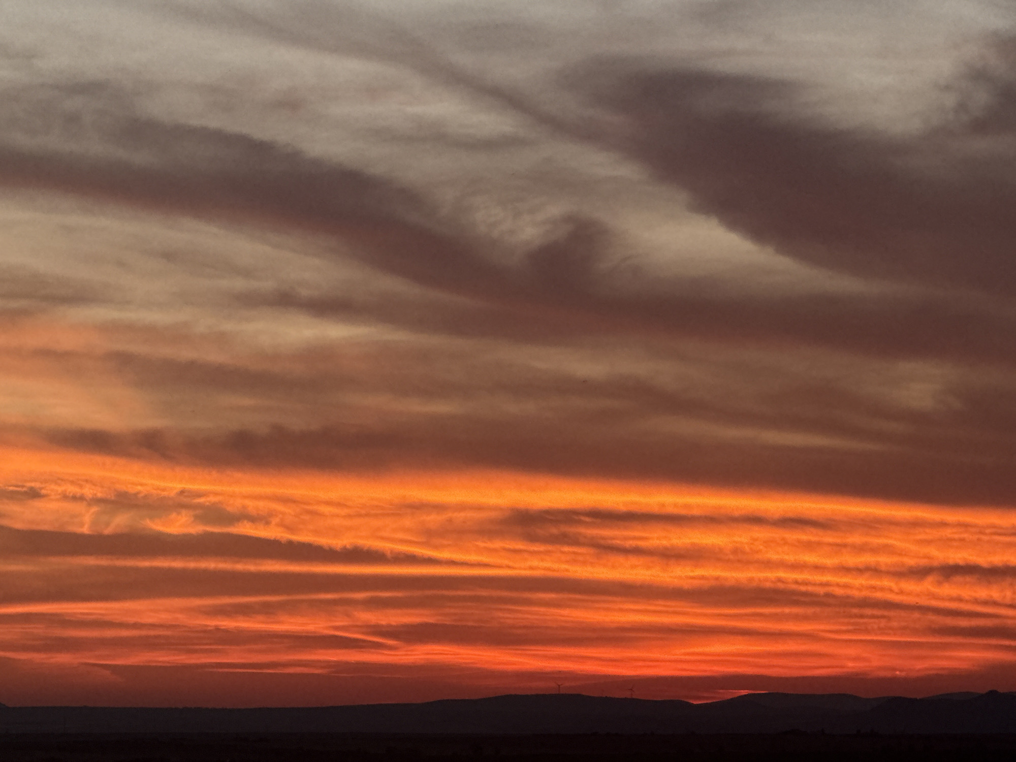 The image depicts a vibrant sunset over the landscape near Burgas, Bulgaria. The sky is filled with a dramatic display of clouds, ranging from deep orange and fiery red at the horizon to softer, muted tones of pink and grey higher up. The clouds are horizontally striated, creating a layered effect that emphasizes the colors. In the distance, silhouetted against the colorful sky, are low, dark hills or mountains. There appear to be a few small wind turbines visible on the distant hills. The foreground is a dark, undefined strip, likely representing the land. The overall scene is serene and atmospheric, characterized by the intense colors of the twilight sky.