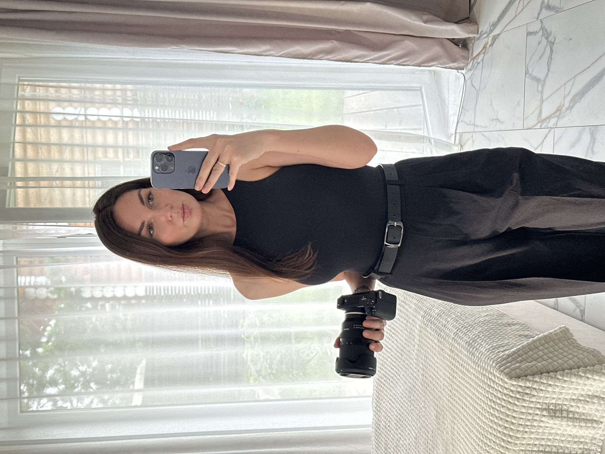 The media file is a vertically oriented, full-body selfie taken in what appears to be a bedroom. The subject is a woman with long, dark hair, holding an iPhone in her left hand to capture the reflection in a mirror. She is wearing a black sleeveless top and dark pants, cinched with a black belt. In her right hand, she holds a black camera with a lens attached.
The background shows a window with blinds and sheer curtains, suggesting it is daytime. Through the window, greenery and trees are visible, indicating an outdoor setting. To the right of the subject, a section of a tiled wall with marble-like patterns and a bed with a textured white cover are visible. The lighting is soft, and the overall mood seems relaxed. There is no visible text in the image. The location context is Chaikovsky, Russia, but this information is not visually deducible from the image itself.