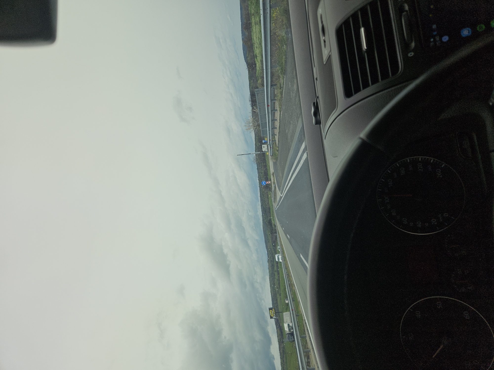 A view from inside a car on a highway, presumably in transit. The car's dashboard, including a speedometer, is visible. The road has white lane markings and is bordered by grassy fields and a guardrail. The sky is overcast. A road sign with a blue circular arrow is visible. The scene appears to be in Dositeevo, Bulgaria, based on the question's premise, though specific identifying landmarks within the village are not apparent in the frame. There are no people visible in the frame.