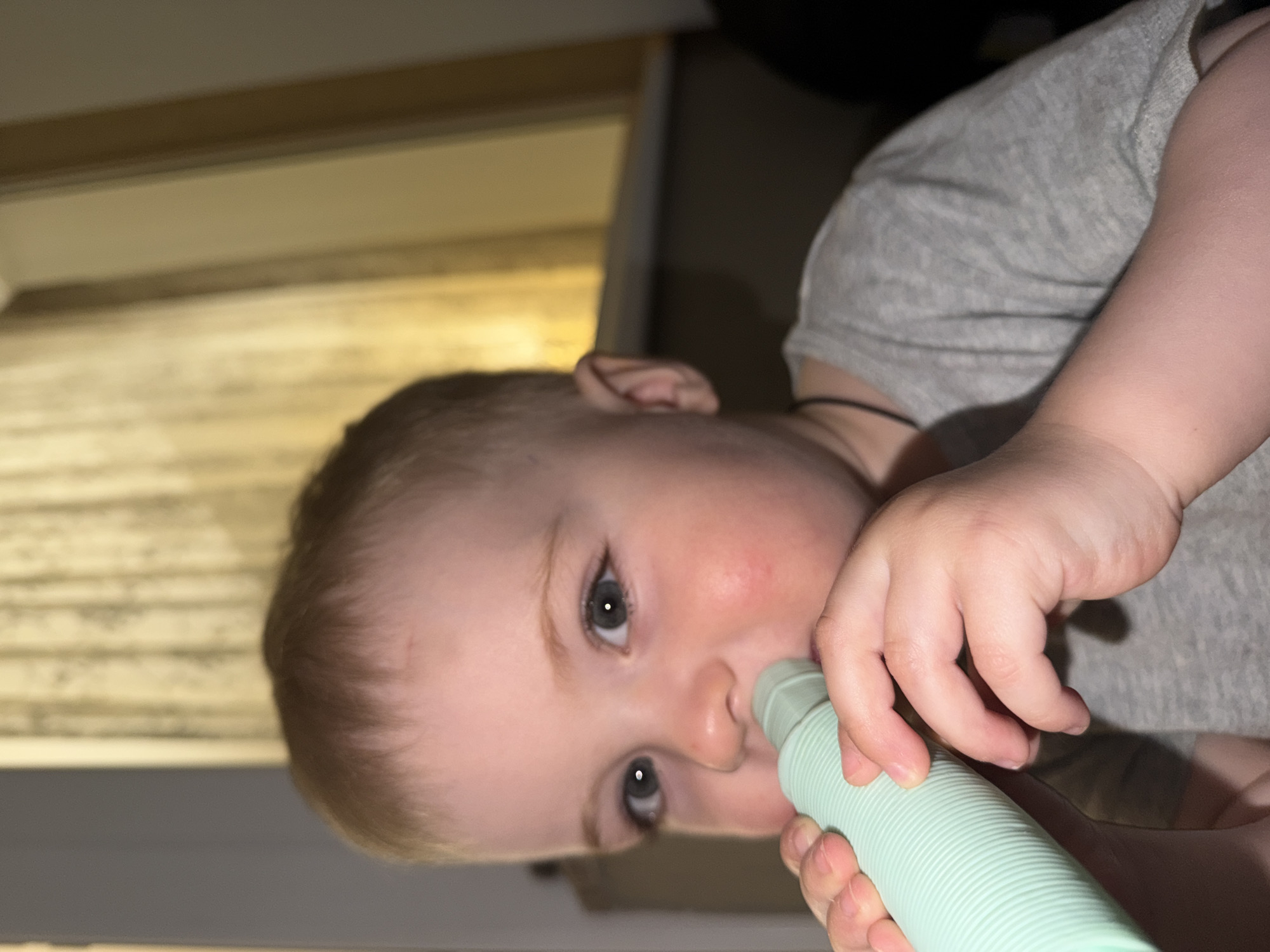 A young child with light skin and short hair is holding a ribbed, light green object to their nose. The child is wearing a grey shirt. The background includes a light-colored wall and a dark object, possibly furniture. The image is rotated, and the location is not identifiable as Votkinsk, Russia.