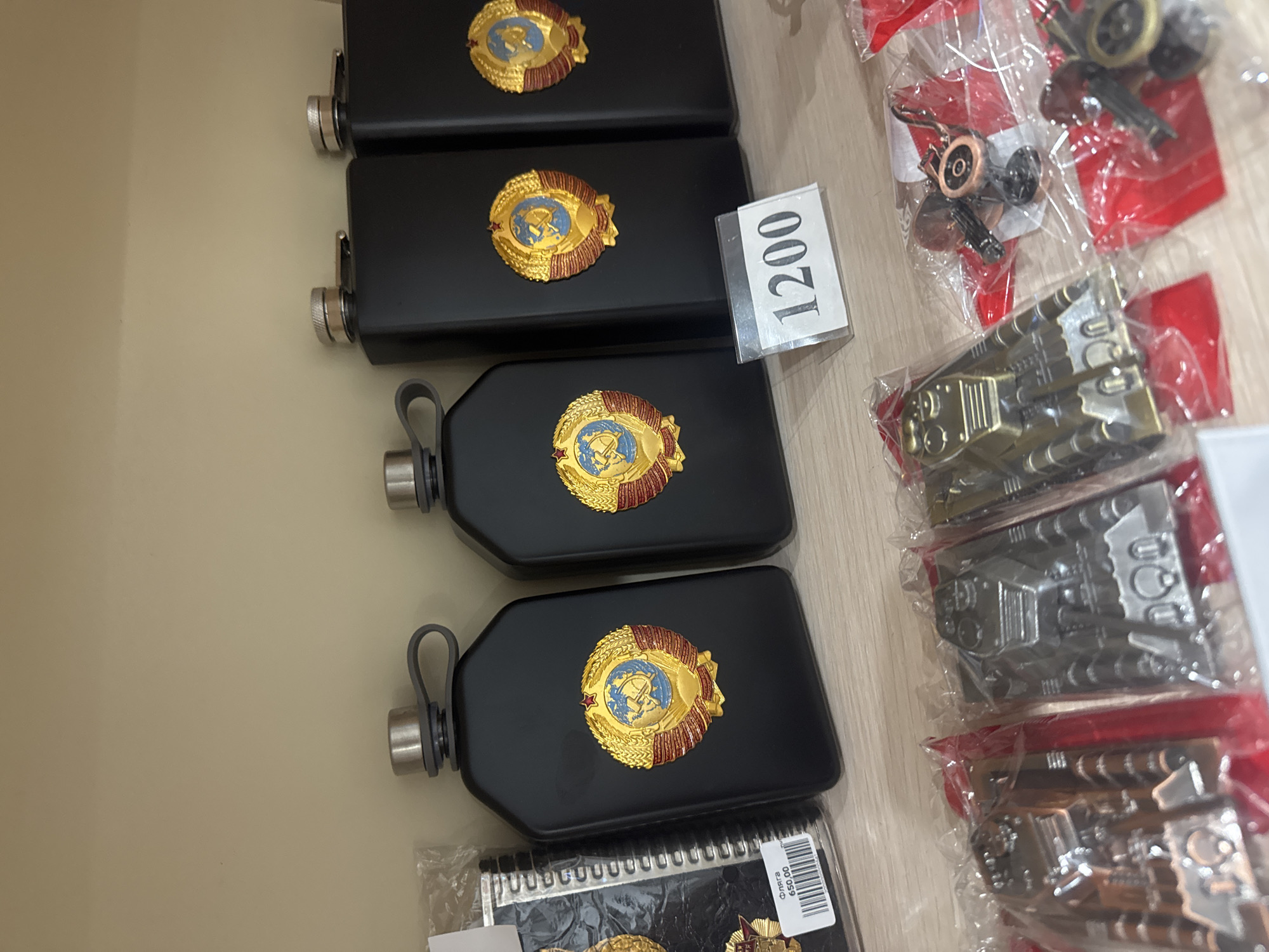 A medium shot captures a shelf displaying several black flasks adorned with a golden Soviet emblem featuring a globe, hammer, sickle, and red star, with red detailing on the outer wreath. Four flasks are visible, three prominently in the foreground and one partially obscured on the far right. A price tag displaying "1200" in black numerals on a white background is placed in front of the third flask from the left. To the left of the flasks, a black notebook with a spiral binding is partially visible, also featuring the Soviet emblem and a price tag that reads "Флага 650.00". In the lower portion of the image, several small, metallic objects, resembling toy tanks and a keychain with a gear-like embellishment, are displayed in clear plastic packaging with red backing. The shelf is made of a light-colored material, and the background wall is a neutral beige.