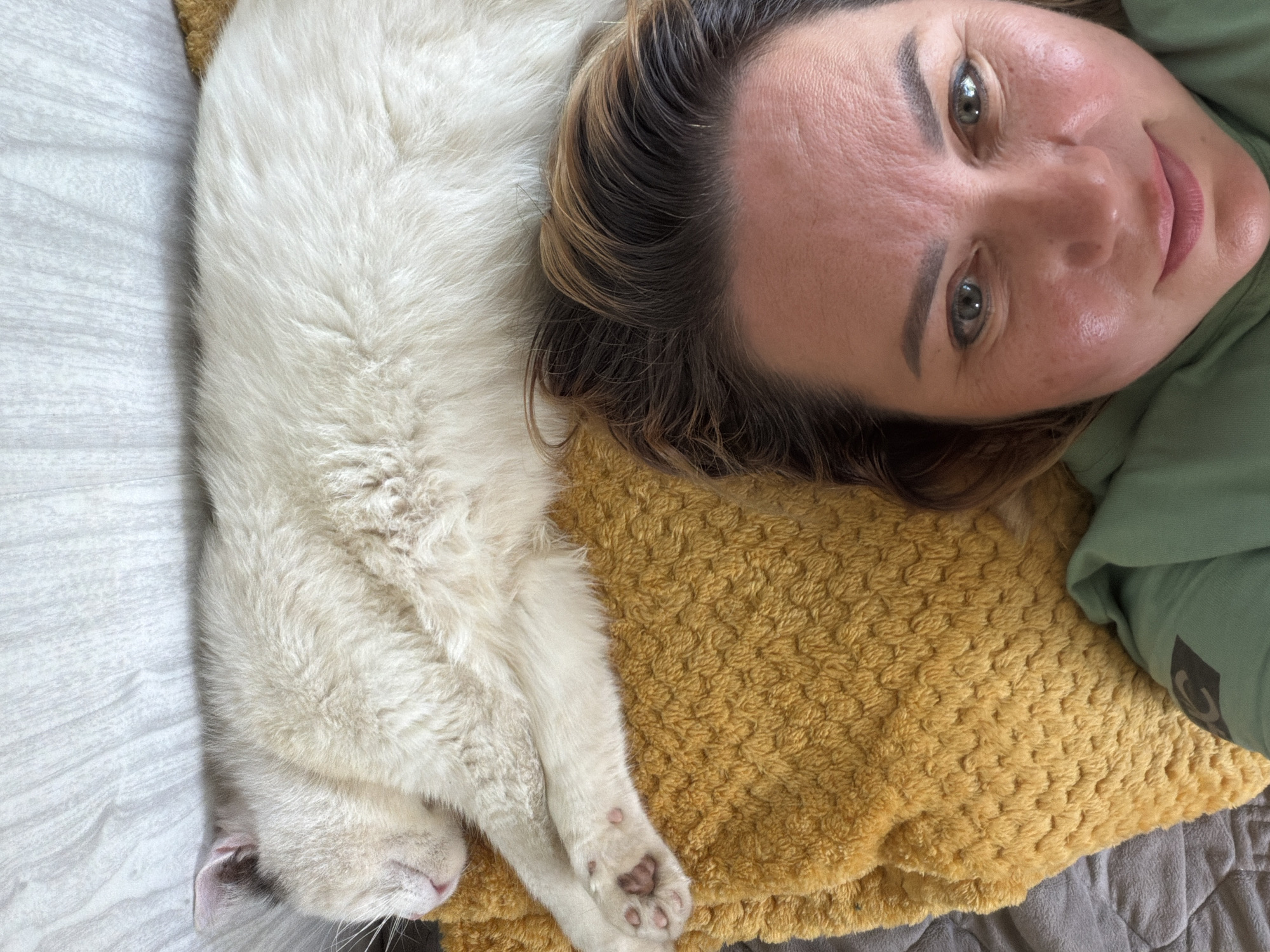 This is a close-up, high-angle selfie of a woman and a white cat lying down together. The woman's head is on the right side of the frame, and her face is angled towards the camera, with her eyes open and a slight smile. She has brown hair with lighter streaks and is wearing a green sweatshirt. The cat is lying next to her, on her left, with its body running horizontally across the frame. It is a fluffy white cat with pink toe beans visible on its paws. The cat's eyes are closed, and it appears to be sleeping or resting.
They are lying on a textured yellow blanket, which is placed on top of a gray, fuzzy blanket or bedspread. To the left of the cat, there is a white surface with a subtle wood grain pattern, possibly a wall or another piece of furniture. The lighting appears to be natural and soft, suggesting it might be daytime. The overall mood of the image is one of comfort and intimacy, with the woman and cat sharing a moment of rest and connection. There are no visible text elements in the image. The location context is Leningradskaya, Russia.