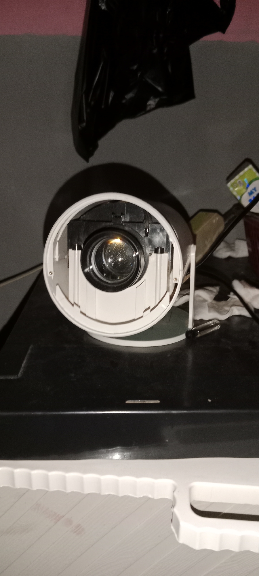 A white projector is placed on a black surface, possibly a table or cabinet. The projector has a visible lens and appears to be part of a larger electronic device. In the background, a black plastic bag hangs from something out of frame. The lighting is dim, creating shadows and a somewhat cluttered environment. There are no people visible in the image. The location is not specified, but the prompt mentions Subang, Indonesia.