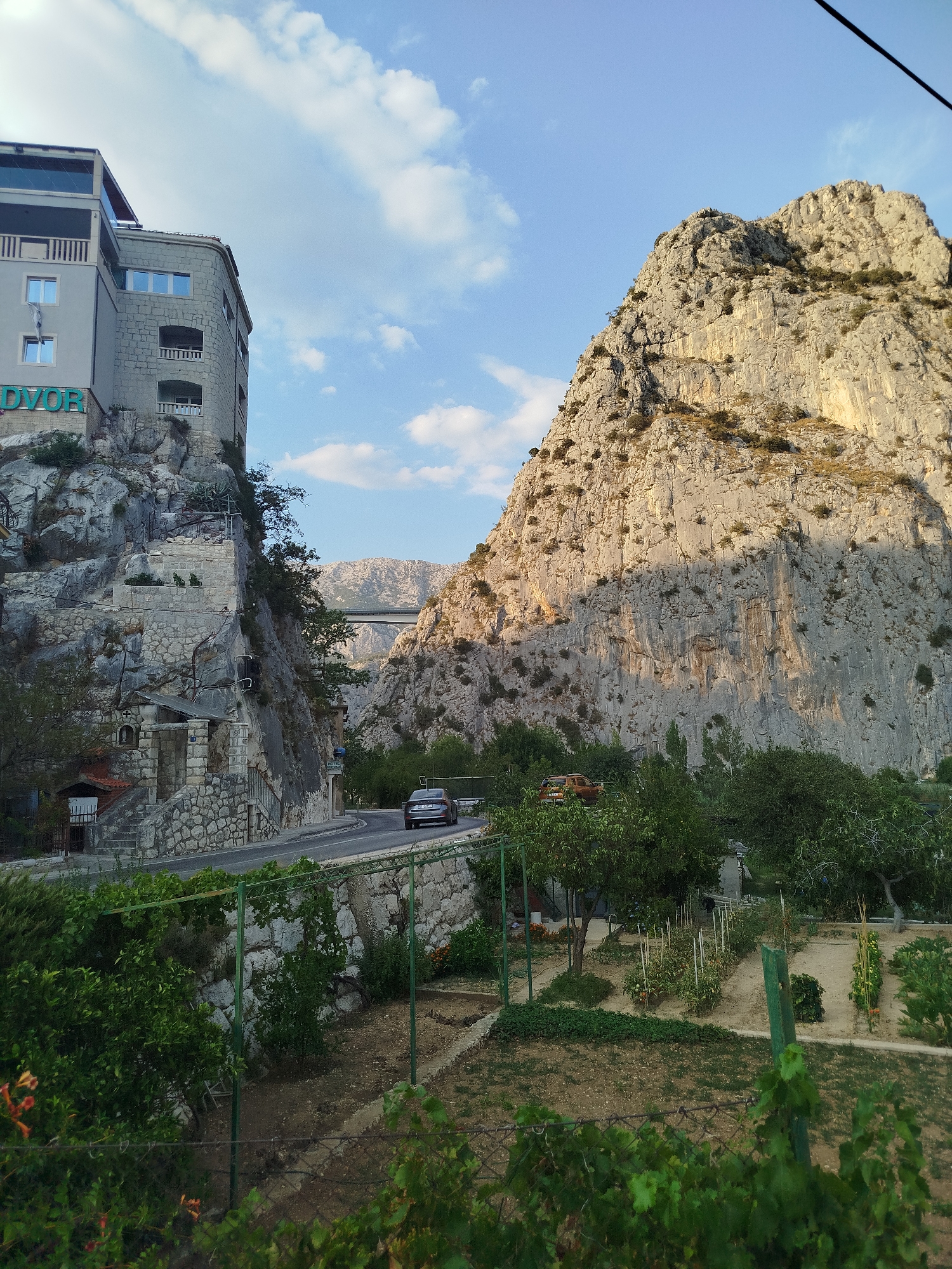 The image depicts a view of the town of Omiš, Croatia, featuring a dramatic landscape where the Cetina River canyon meets the Adriatic Sea. On the left, a stone building, possibly a hotel, is built directly into the side of a steep, rocky cliff. The building has multiple stories, balconies, and windows, with the word "DVOR" visible in large, teal letters near the top. A paved road winds its way up the cliff, with a silver sedan driving on it.
Dominating the right side of the image is a massive, sheer rock face that is characteristic of the Omiš area. This rock formation is rugged and textured, with sparse vegetation clinging to its surfaces. The setting sun casts long shadows across the rock, highlighting its dramatic contours.
In the foreground, a garden area is visible, with rows of plants, some supported by stakes. Green metal fencing encloses parts of the garden. Trees and bushes are interspersed throughout the scene. In the distance, a bridge spans across the canyon, and further beyond, hazy mountains can be seen. The sky above is a clear blue with scattered white clouds, suggesting it is late afternoon or early evening. The overall impression is one of a striking juxtaposition between human architecture and the imposing natural environment of Omiš.