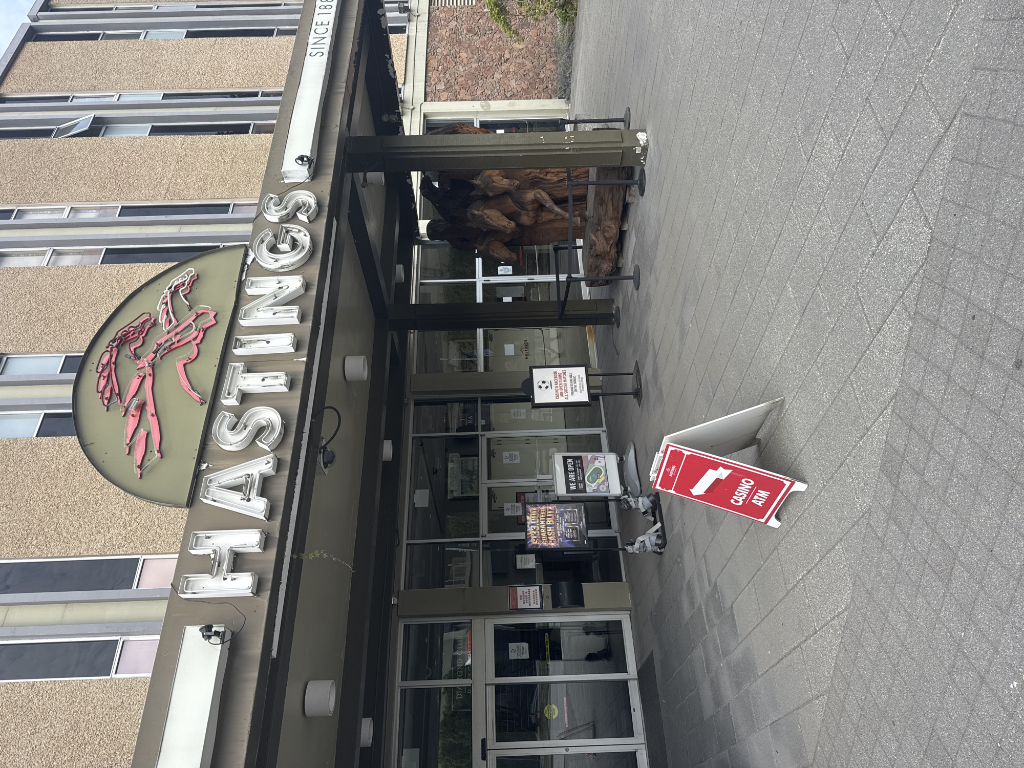 The exterior entrance to the Hastings Racecourse Casino in Vancouver, Canada, is depicted. A large, illuminated sign above the entrance reads "HASTINGS" in white block letters, with a neon horse racing motif above it. To the right of the sign, the text "SINCE 1889" is visible. The building's facade is textured concrete, with numerous windows and a dark overhang covering the entrance.
In front of the entrance, a large wooden sculpture of several horses stands on a platform. To the left of the sculpture, glass doors lead into the casino. Various signs are posted near the entrance, including one for the "DIAMOND CLUB" and a red A-frame sign that reads "CASINO ATM" with an arrow pointing to the right. In the background, the architecture of the building suggests a mid-20th-century construction style. The ground is paved with gray interlocking tiles. No people are clearly visible in the image.