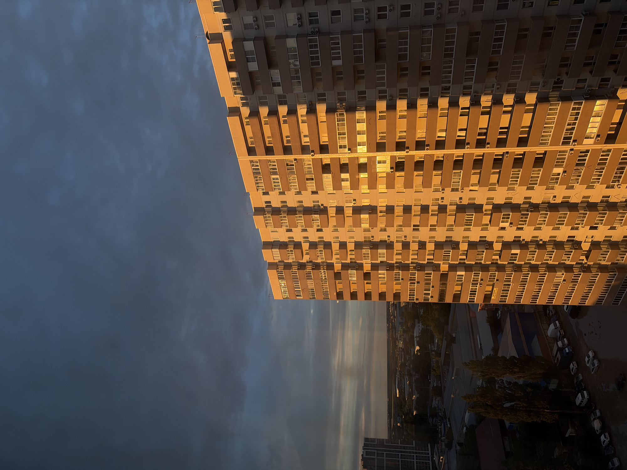 The image captures a dramatic urban scene at either sunrise or sunset, dominated by a tall, multi-story residential or office building. The building, positioned on the right side of the frame, is intensely illuminated by warm, golden light from a low sun, highlighting its numerous rectangular windows, balconies, and air conditioning units. Its facade features a repeating pattern of light beige and darker brown sections.
In stark contrast, the left side of the image is filled with an overcast, dark blue-grey sky, hinting at cloud cover. Some lighter streaks are visible within the clouds towards the horizon, suggesting the sun might be breaking through or reflecting off the cloud base. Below the main building, a shadowed cityscape extends, revealing other buildings, trees, and a parking area with several parked cars, all largely in deep shadow. No people are visible. The strong interplay of light and shadow creates a striking and moody atmosphere within this generic urban setting. No discernible text is visible.