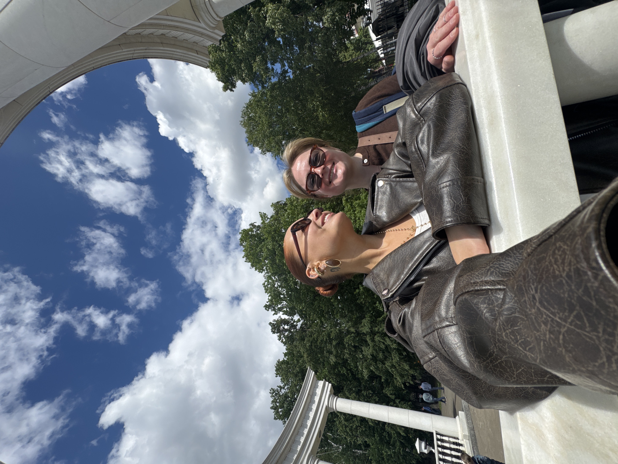Two women are posing for a photo under an arch. The woman on the left has auburn hair pulled back and is wearing a brown leather jacket and sunglasses. The woman on the right has blonde hair and is also wearing sunglasses and a brown jacket. They are leaning against a white marble structure. In the background, there are green trees and a blue sky with white clouds. The location is not identifiable as Izhevsk, Russia based solely on the provided image.