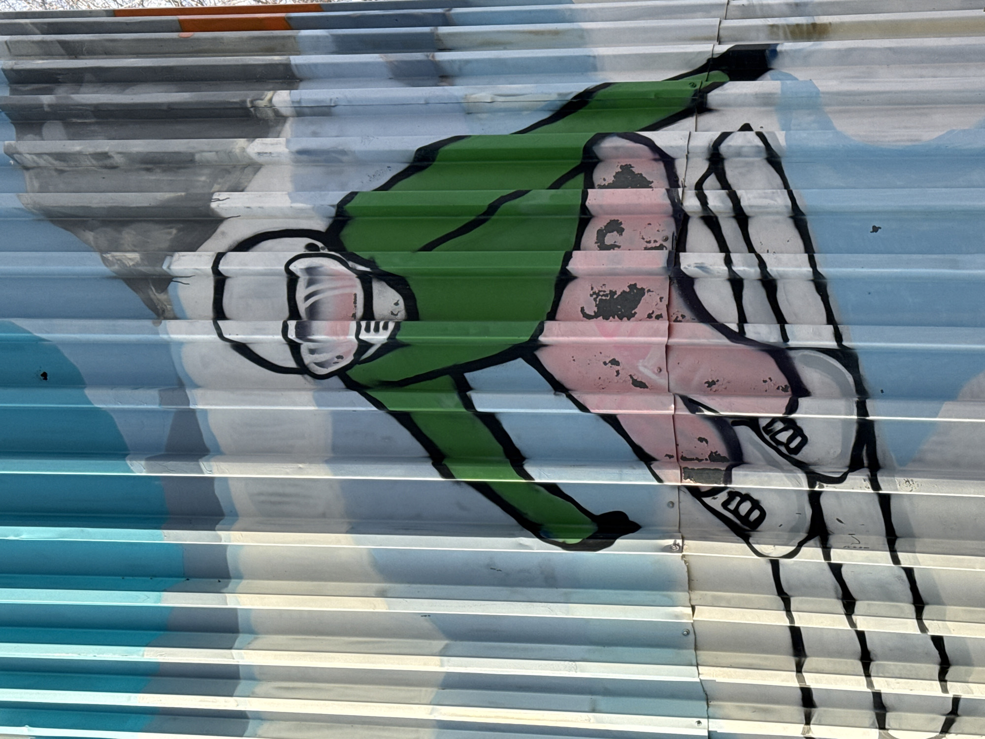 The image shows a graffiti art mural on a corrugated metal wall. The mural depicts a figure with a large head, wearing what appears to be a green jumpsuit, and pink skin. The figure is in a leaning or falling position. The mural incorporates clouds in the background, with hues of light blue, grey, and white. Black outlines are used to define the figure and the clouds. The style is cartoonish and simplified. The location, Galevo, Russia, is not explicitly visible within the frame.