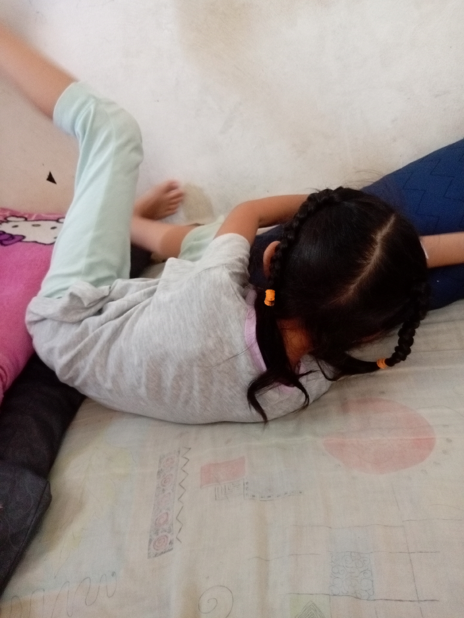 This media file shows a young child lying on a bed. The child has dark hair styled in two braids with orange hair ties. They are wearing a light gray t-shirt and light blue pants. The child's body is positioned in a way that suggests they may be stretching or in a playful pose.
The setting appears to be a bedroom. The bed has a light-colored sheet with a subtle patterned design. To the left of the child, there is a pink pillow with a Hello Kitty pattern. Behind the child, a dark blue pillow or cushion is visible. The background wall is a plain, light color.
The lighting suggests it could be daytime, possibly indoors with natural light. There are no clear indications of the time of day or weather conditions. The child's posture and the overall scene do not convey any specific emotions beyond what might be considered a relaxed or playful state. No text is visible in the image. The location context provided is Batam, Indonesia.