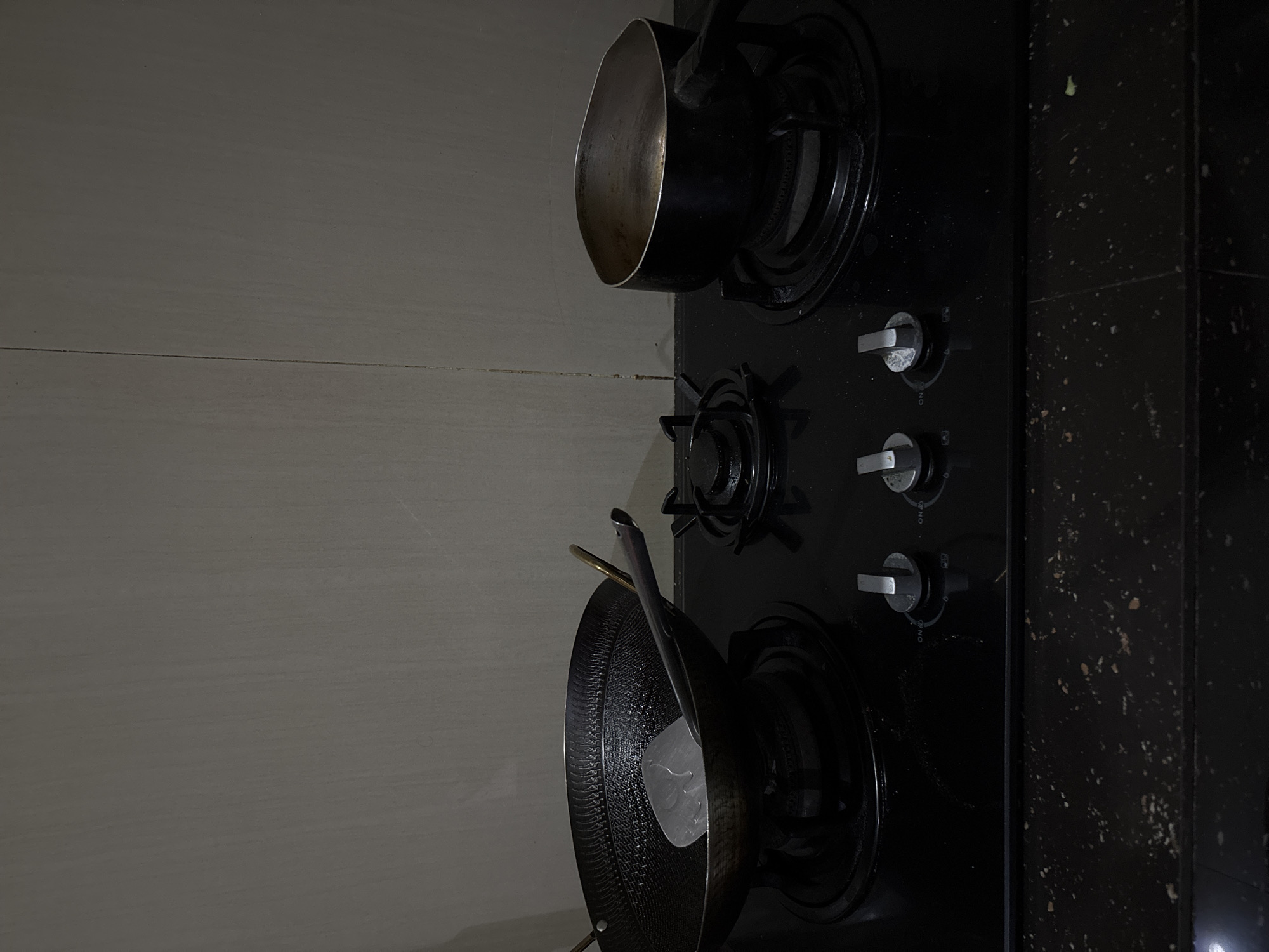 The image depicts a kitchen stovetop with a pot and a wok. The stovetop is black and has three burners with silver control knobs. A silver pot sits on the top right burner, and a black wok with a metal spatula is on the bottom right burner. The stovetop is positioned next to a tiled wall. The environment suggests a domestic kitchen setting. The image was taken in Binong, Indonesia.