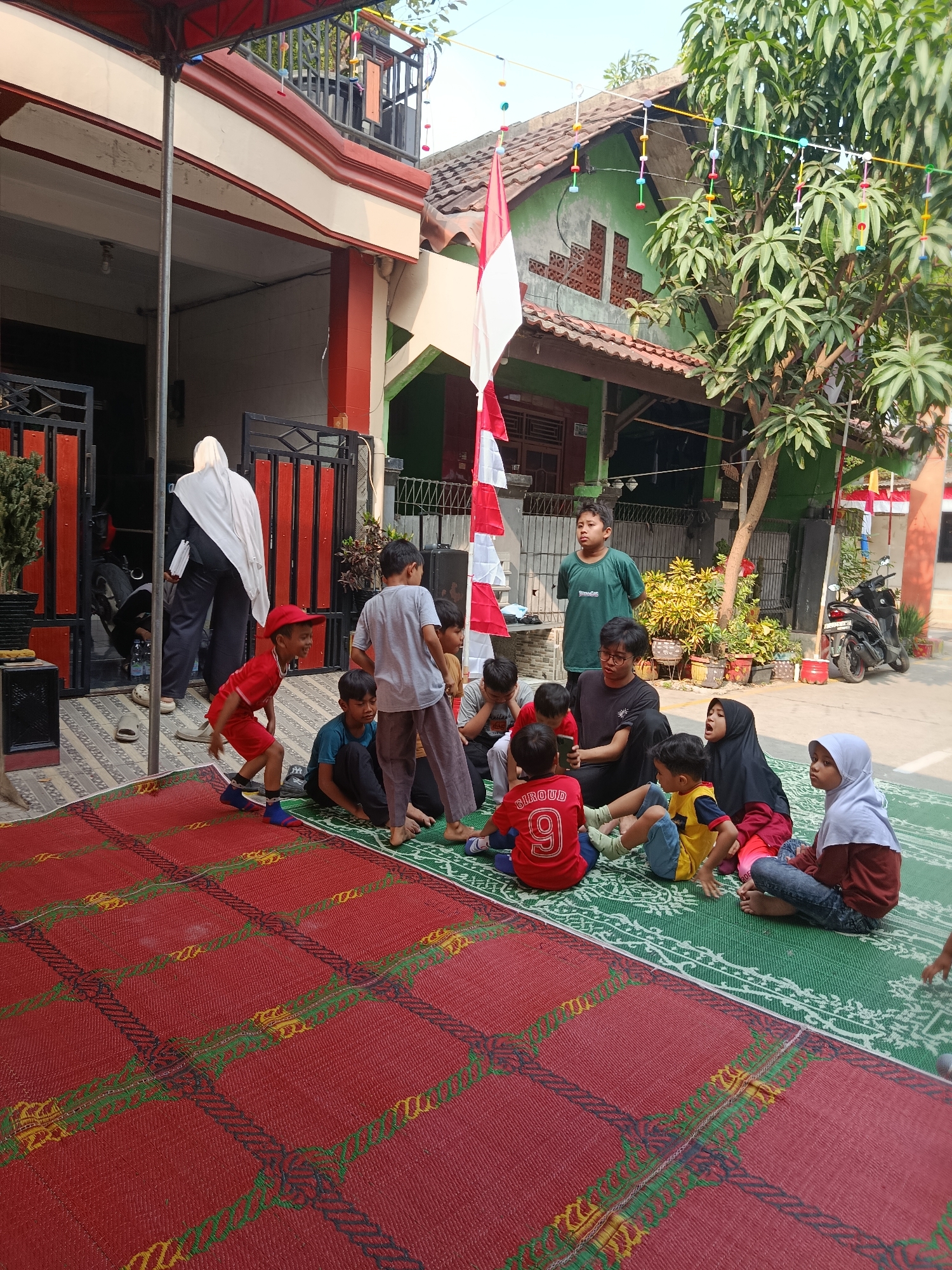 In Wanasari, Indonesia, a group of children is gathered on mats outside a building. The scene appears to be a gathering or event, possibly a school or community activity, judging by the presence of children of varying ages and the festive decorations.
The foreground is dominated by a large red mat with a black, red, and green checkered pattern, featuring rope-like designs in yellow and green. To the right of the red mat is a green mat with a white intricate pattern. Several children are sitting on these mats, facing inwards, suggesting they are engaged in a shared activity or listening to someone.
In the mid-ground, a cluster of children are sitting and interacting. One child, wearing a red shirt with the number '9' and the name 'GIROUD' on the back, is facing away from the camera. Another child in a yellow and blue shirt is sitting next to them. A child in a black hijab and a pink shirt is also present, looking towards the other children. A young child in a light blue shirt and jeans is sitting on the green mat, looking to the right.
Further back, a group of boys are standing and sitting. One boy in a green t-shirt is standing and looking forward. Another boy in a dark t-shirt is sitting with others. A few more children are seated around them, including one in a red cap.
Behind the children, a woman in a white hijab and dark clothing is walking away from the camera, carrying some items. She is near an entrance with a black gate. The building itself is a single-story structure with red and white accents and a green roof. A national flag of Indonesia (red and white) is displayed prominently on a pole. Decorative colorful strings with small circular objects are strung across the scene, adding to the festive atmosphere.
To the right of the building, a motorcycle is parked. The environment suggests a residential or community outdoor space, with a tree providing some shade. The ground is paved. The overall impression is a lively and communal gathering.