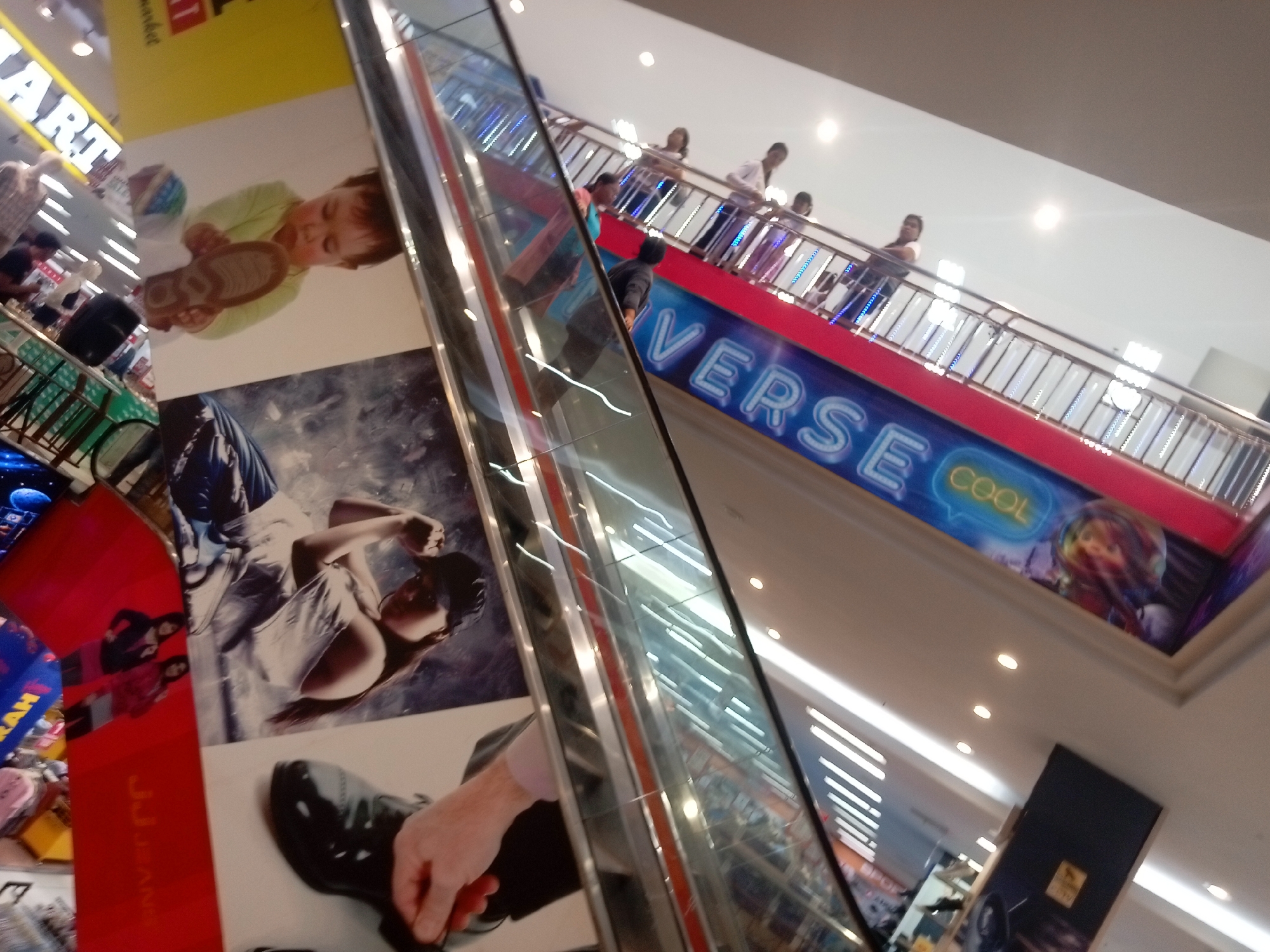 The image shows an escalator in a mall in Batam, Indonesia. The escalator has a red trim and a blue banner with the word "VERSE" in white letters and "COOL" in yellow letters. People are seen riding the escalator. On the walls of the mall, there are large advertisements. One advertisement shows a baby holding a shoe, and another shows a woman. The lighting in the mall is bright, with overhead lights visible. The overall environment appears to be a busy commercial space.