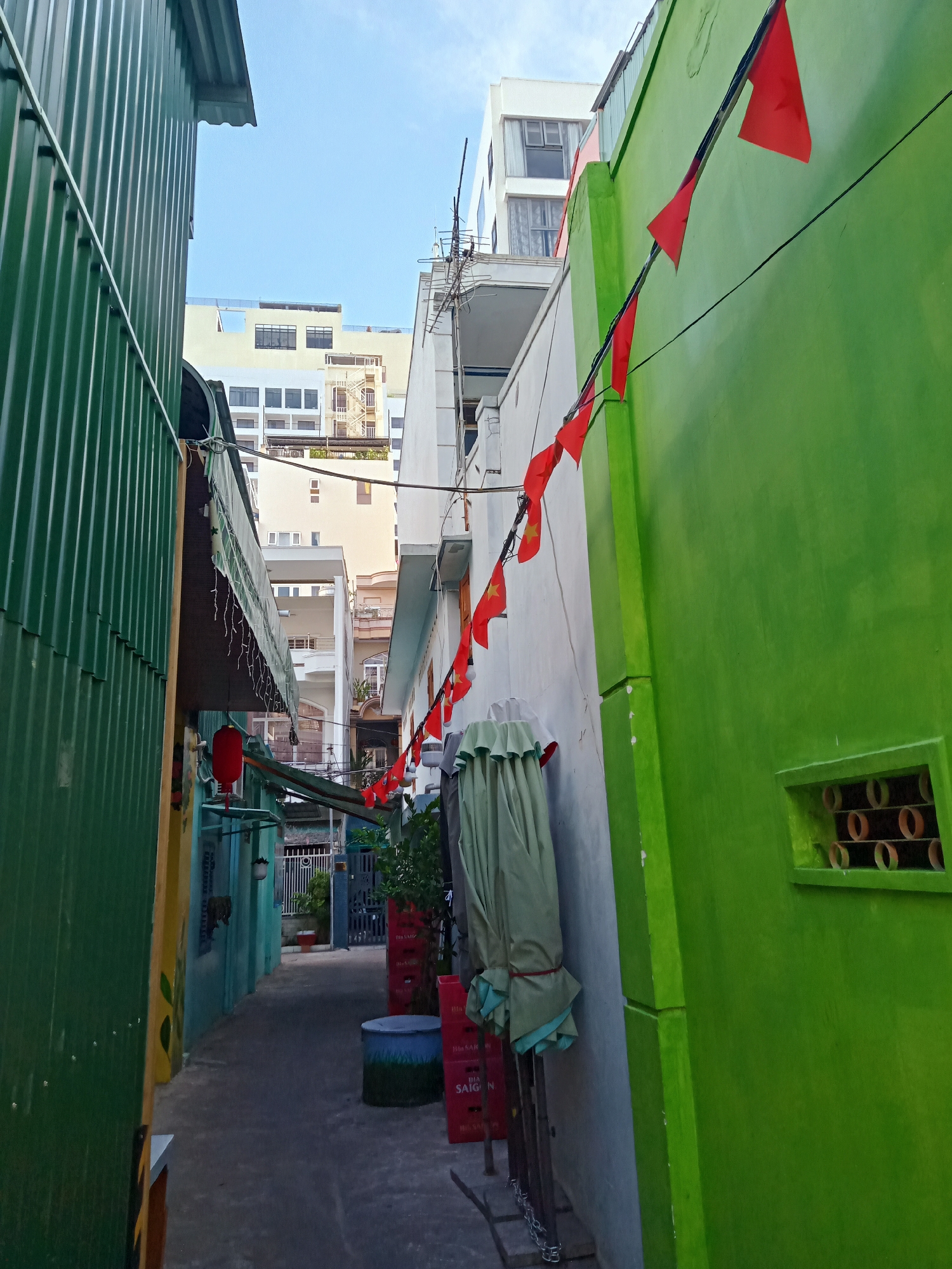 A narrow alley in Nha Trang Ward, Vietnam, is lined with buildings of varying heights and colors, including a corrugated green structure on the left and a bright green wall on the right. Red triangular flags are strung along the alley, suggesting a possible celebration or patriotic display. A covered object leans against a wall, next to stacked cardboard boxes. A small opening in the green wall reveals a series of round shapes, possibly vents or decorative elements. Overhead, the sky is visible between the buildings.