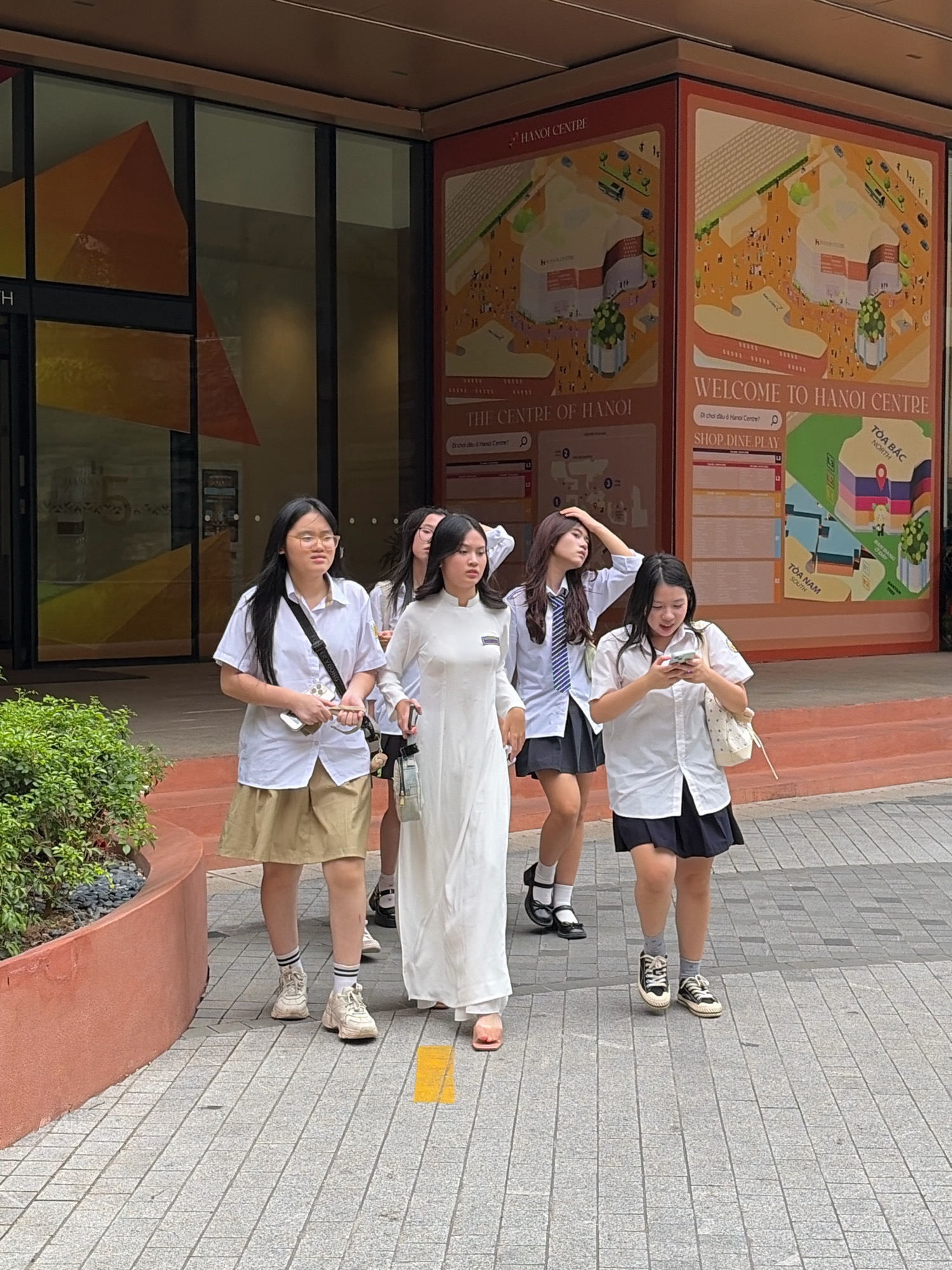 A group of five young women, dressed in school uniforms, walk on a paved walkway in front of a building. The woman in the front left is wearing a white shirt, tan skirt, and sneakers. She has black hair and glasses. To her right, a woman in a white dress walks ahead. Behind them, a woman in a white shirt, blue tie, and black skirt walks with her hand on her head. To her right, a woman in a white shirt and black skirt looks down at her phone. Another woman, partially obscured, walks behind the first woman. The building behind them has large windows with a graphic featuring a large orange geometric shape. A large poster is also visible, with text in English and Vietnamese, including "WELCOME TO HANOI CENTRE" and what appears to be a map of the shopping center. The walkway is made of gray pavers with yellow markings. Green foliage is visible to the left. The overall scene suggests they are in a commercial or educational district within Hanoi, Vietnam.