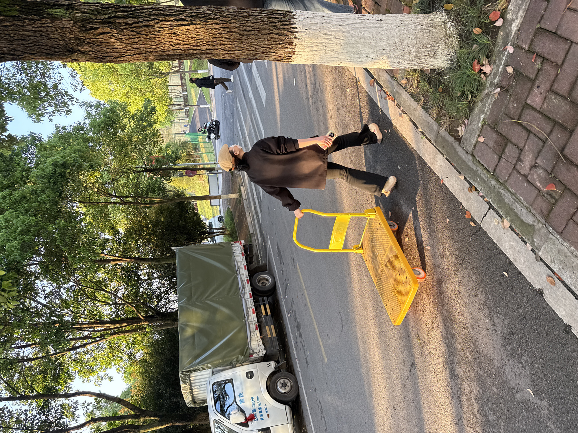 A person wearing a hat, a black jacket, and black pants is walking on a street while pulling a yellow handcart with a platform. The person is also holding a phone. A white truck with a green cover is parked on the side of the street. Trees line the street. The location is Jiangjin, China. Other people and vehicles are in the background.