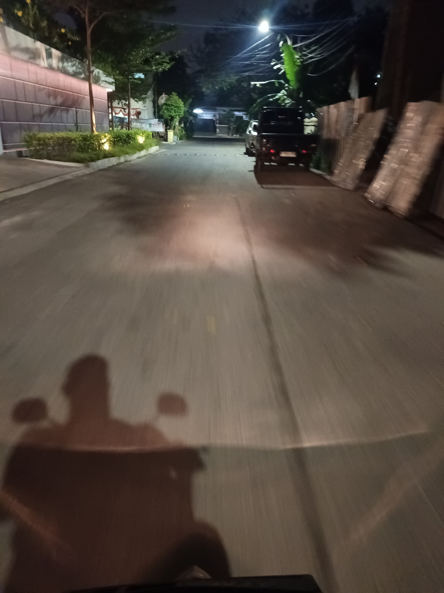 The image shows a street scene at night in North Jakarta, Indonesia, likely in a residential or commercial area. The perspective is from the viewpoint of someone riding a motorcycle, indicated by the shadow of a rider and handlebars in the foreground. The road surface is asphalt, with visible lane markings and reflections from a light source, possibly a street lamp or the motorcycle's headlight, illuminating the road ahead.
On the left side of the street, there is a building with a textured wall and illuminated landscaping, featuring trimmed bushes and small trees. Further down the street, a black pickup truck is parked, and there are some stacked materials that appear to be corrugated metal or similar sheeting on the right side of the road. The environment is dark, with a few lights visible, suggesting it's late at night. The overall impression is a mundane street view, captured during transit.