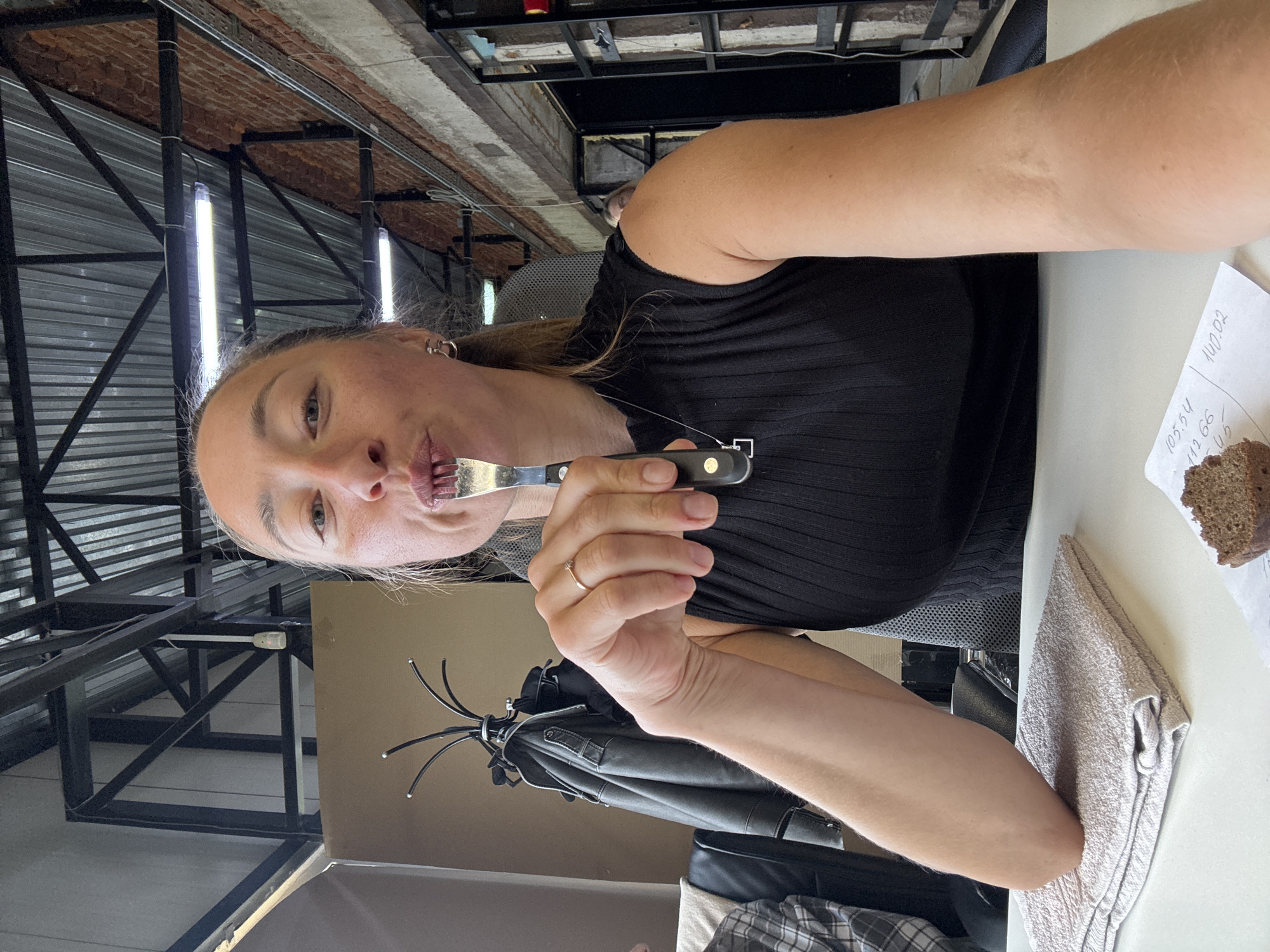A woman is at a table in a room with an industrial-style ceiling that features metal beams and hanging light fixtures. She holds a fork to her mouth. On the table are a towel, a slice of bread, and a piece of paper with handwritten notes. A coat rack with a jacket is visible in the background. The location is implied to be Chaikovsky, Russia.