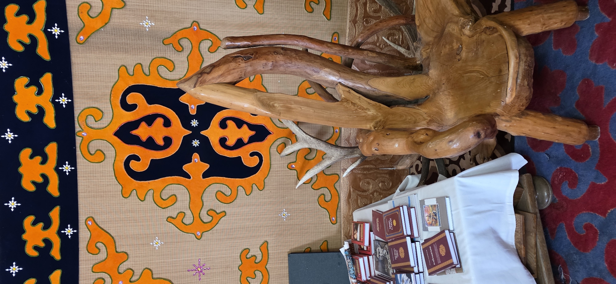 In the city of Bulan-Segettu, Kyrgyzstan, an interior scene is captured, likely within a traditional dwelling. The background features a textile wall hanging with a dark blue border adorned with an intricate orange pattern resembling stylized floral or tribal motifs. Small white star-shaped embellishments are scattered across the textile.
In the foreground, a table draped with a white tablecloth is laden with a collection of books, predominantly red-bound with gold lettering. To the right, a striking chair carved from natural wood, featuring gnarled branches and a unique, organic shape, dominates the scene. Antlers are visible emerging from behind the chair. The floor is covered by a rug with a dark blue background and a repeating red, swirling pattern. A dark rectangular object, possibly a folder or book, rests on the table to the left. The overall impression is one of cultural display and rustic artistry.