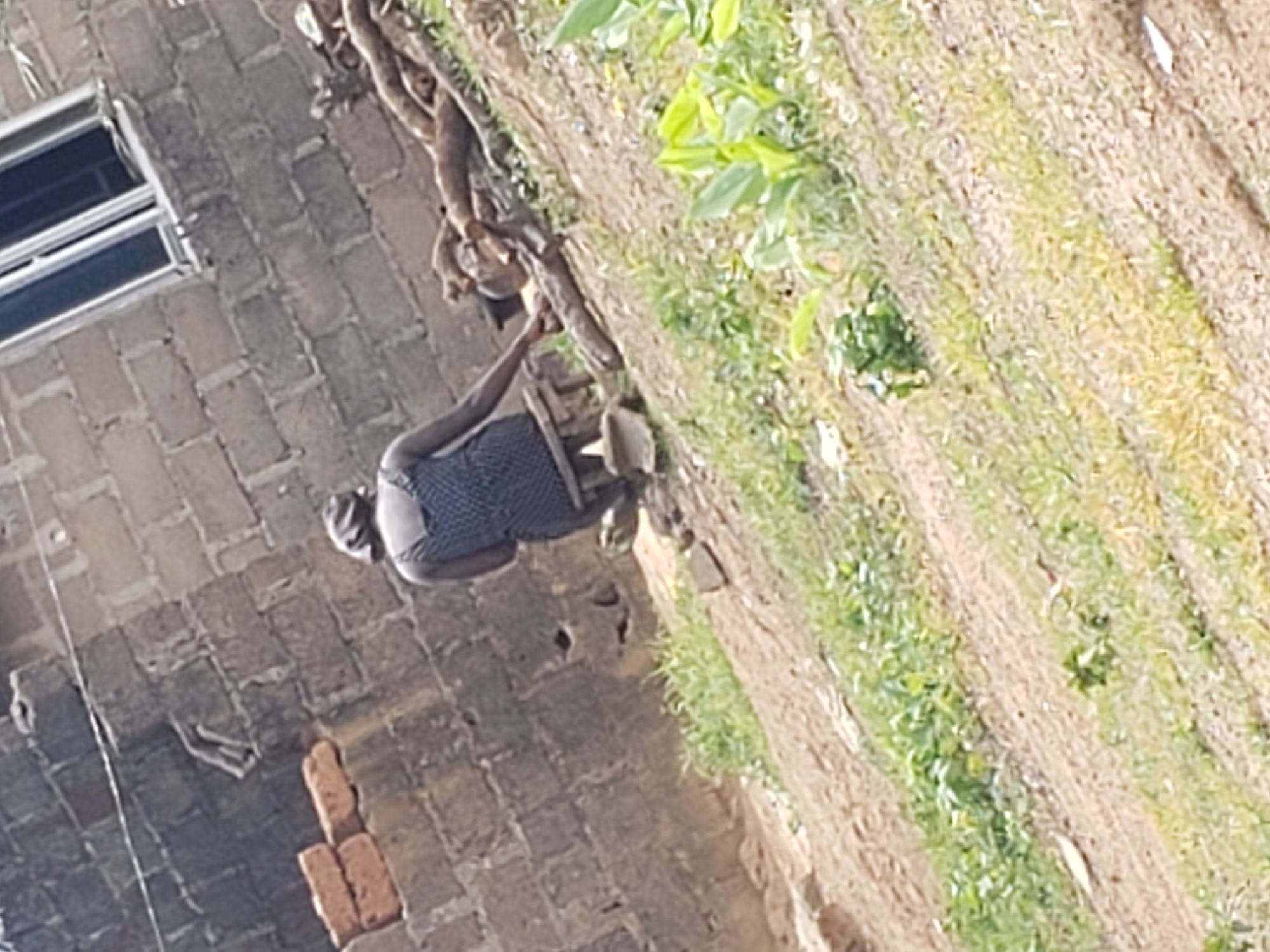 In Kachia, Nigeria, a person sits on a low stool outside a brick building, facing away from the camera. The person is wearing a dark, patterned garment. Next to the person, a pile of logs or branches is visible. The ground is a slope covered in dirt and sparse vegetation. A portion of the brick building, featuring a window, is visible in the background.