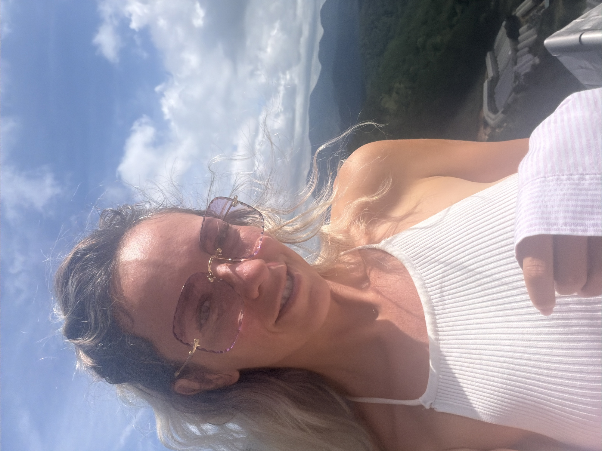 In this close-up selfie, a woman with blonde hair is pictured against a backdrop of a cloudy blue sky and lush green mountains. She is wearing large, pink-tinted sunglasses and a white ribbed halter top. Her hair is windswept, framing her face as she smiles directly at the camera. Her right hand is visible in the lower portion of the image, holding a piece of fabric with purple and white stripes. The background suggests a mountainous or elevated location, possibly overlooking a valley or settlement. The image appears to be taken outdoors on a bright, possibly windy day. While the prompt requests specific location details in Xuan Truong - Da Lat, Vietnam, these are not visually discernible from the provided image itself.