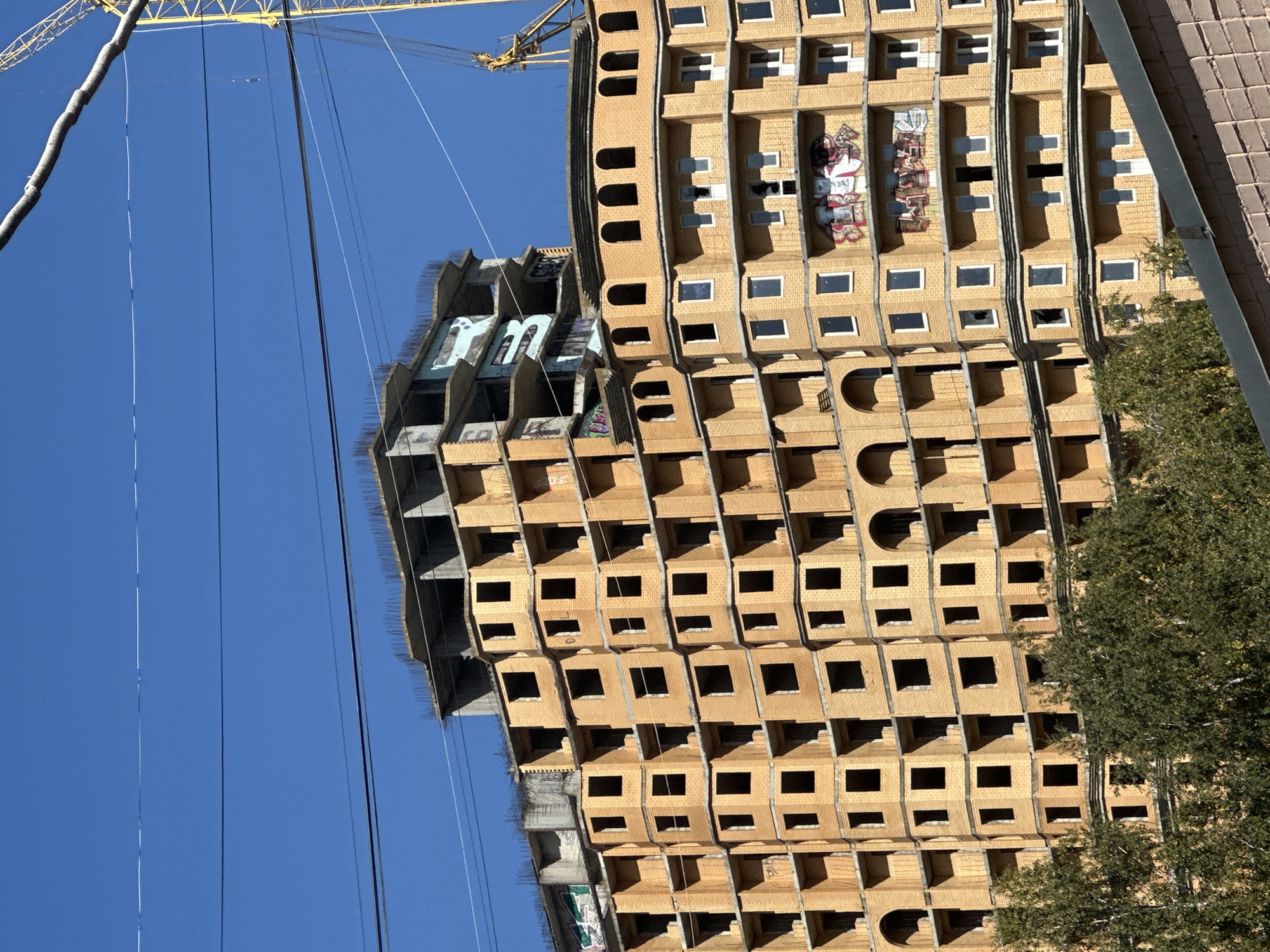 The image shows a large, unfinished building under construction in Podolsk, Russia. The building is made of light brown bricks and has many windows, most of which are empty openings. The upper floors of the building are still under construction, with exposed rebar and concrete. There is graffiti on some of the walls. A yellow construction crane is visible in the background, along with power lines overhead. Trees and foliage are in the foreground, suggesting the building is in a developed area. The sky is clear and blue.