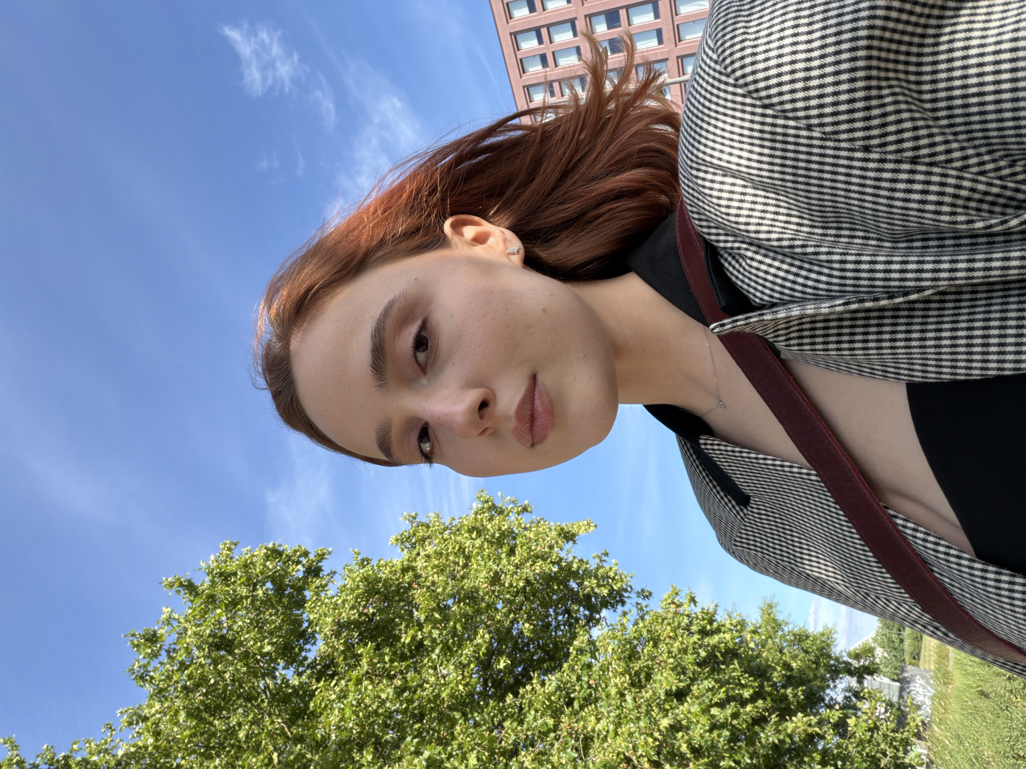 A young woman with red hair is shown from the chest up, looking directly at the camera in a selfie. She is wearing a black top with a blazer over it that has a black and white checkered pattern. A maroon strap is visible across her chest. Her hair is blowing in the wind. In the background, a large green tree is on the left, and a pink building with many windows is on the right. The sky is blue with a few white clouds. The location is the city of Strasbourg, France.