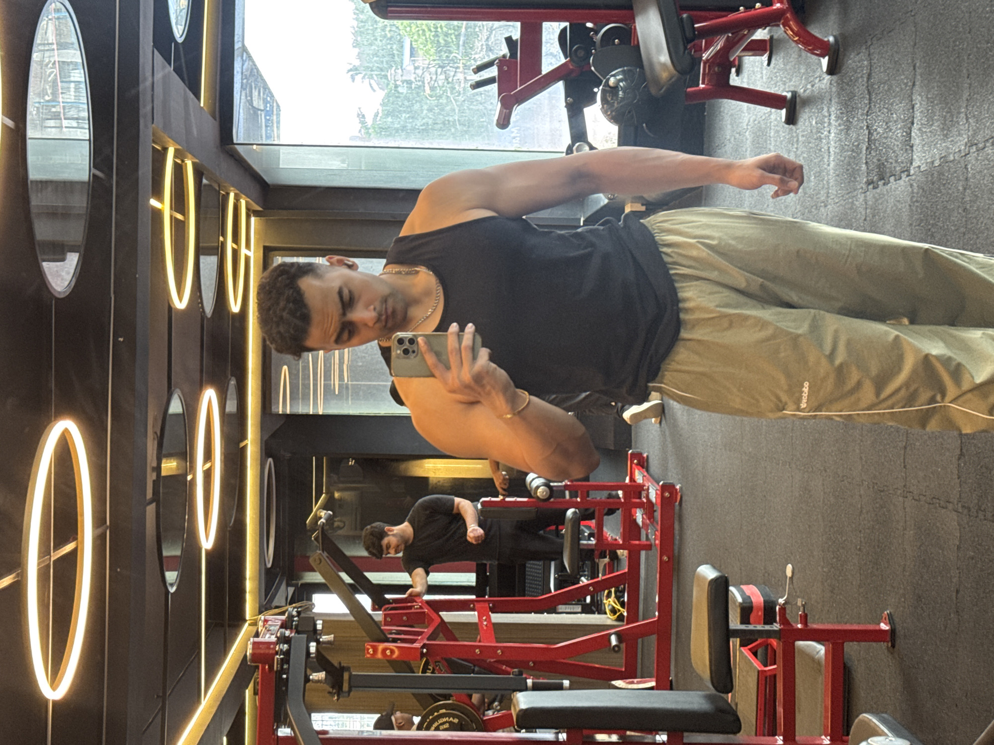 A man is taking a selfie in a gym. He is wearing a black tank top and khaki pants. The gym has red weight machines and a dark ceiling with circular lights. In the background, another man is using a weight machine. The gym appears to be in an urban setting, with windows showing trees and buildings. This image does not provide enough information to determine if the location is Kolkata, India.