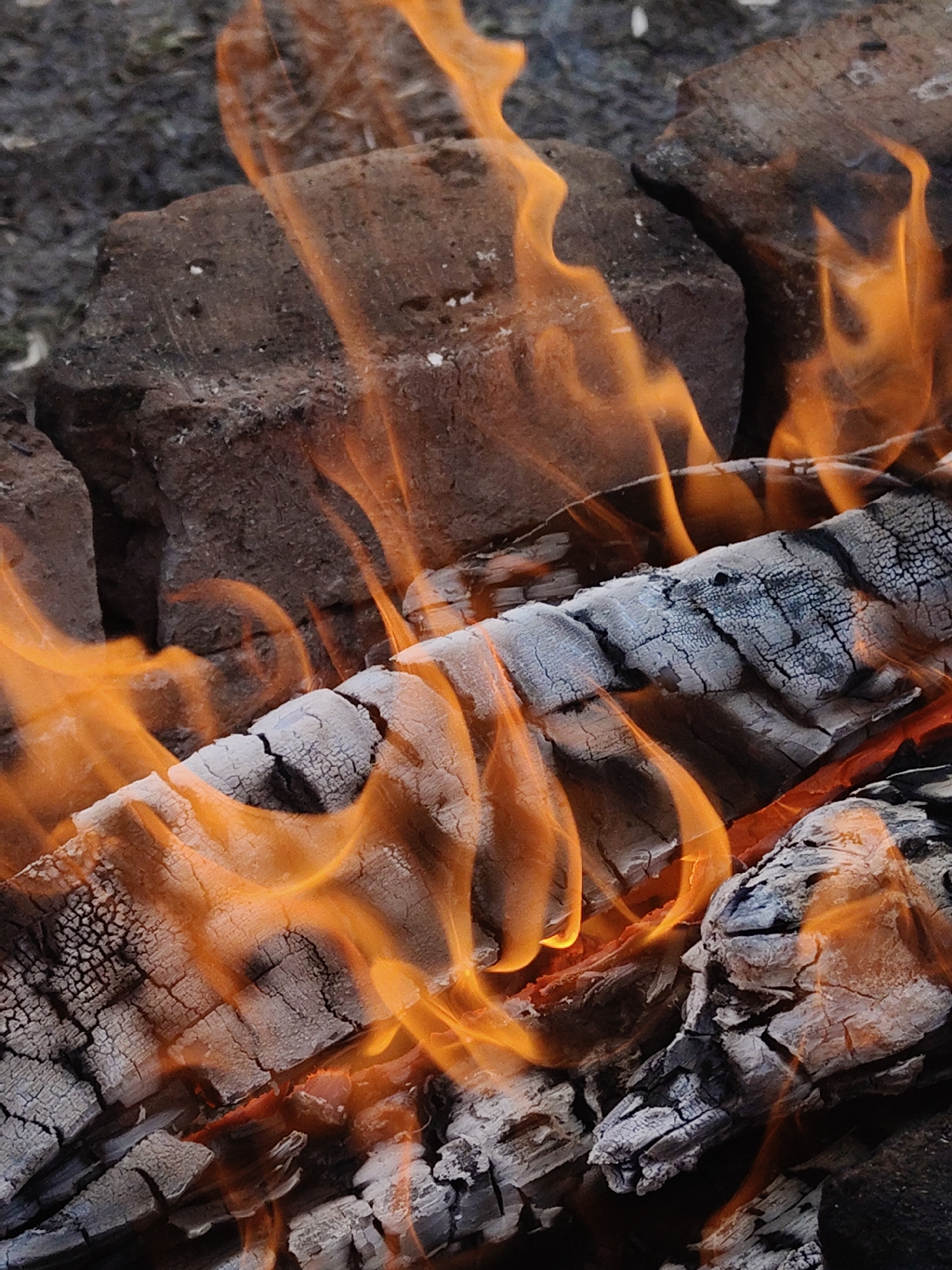 The image depicts a close-up view of a fire. Visible elements include burning wood and flames. The wood appears charred and cracked, indicating prior burning. The flames are orange and yellow. A small brick arrangement is visible in the background. The location is Unknown, Unknown.
