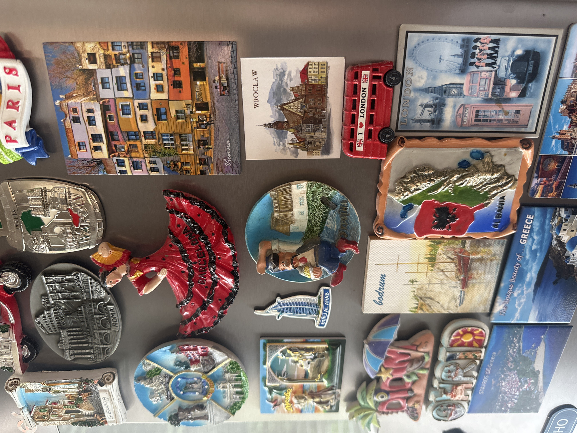 The image shows a collection of magnets on a refrigerator. The magnets depict various cities and countries, including Paris, Vienna, Wrocław, London, Albania, Greece, Bodrum, Djerba, Genoa, Rome, Dubai (Burj Al Arab), and Stavros in Greece. The magnets are of different shapes and sizes, with some being rectangular, round, or three-dimensional. Some magnets feature landmarks, buildings, or iconic symbols associated with the respective locations. There is no indication of Sofia, Bulgaria in the image.