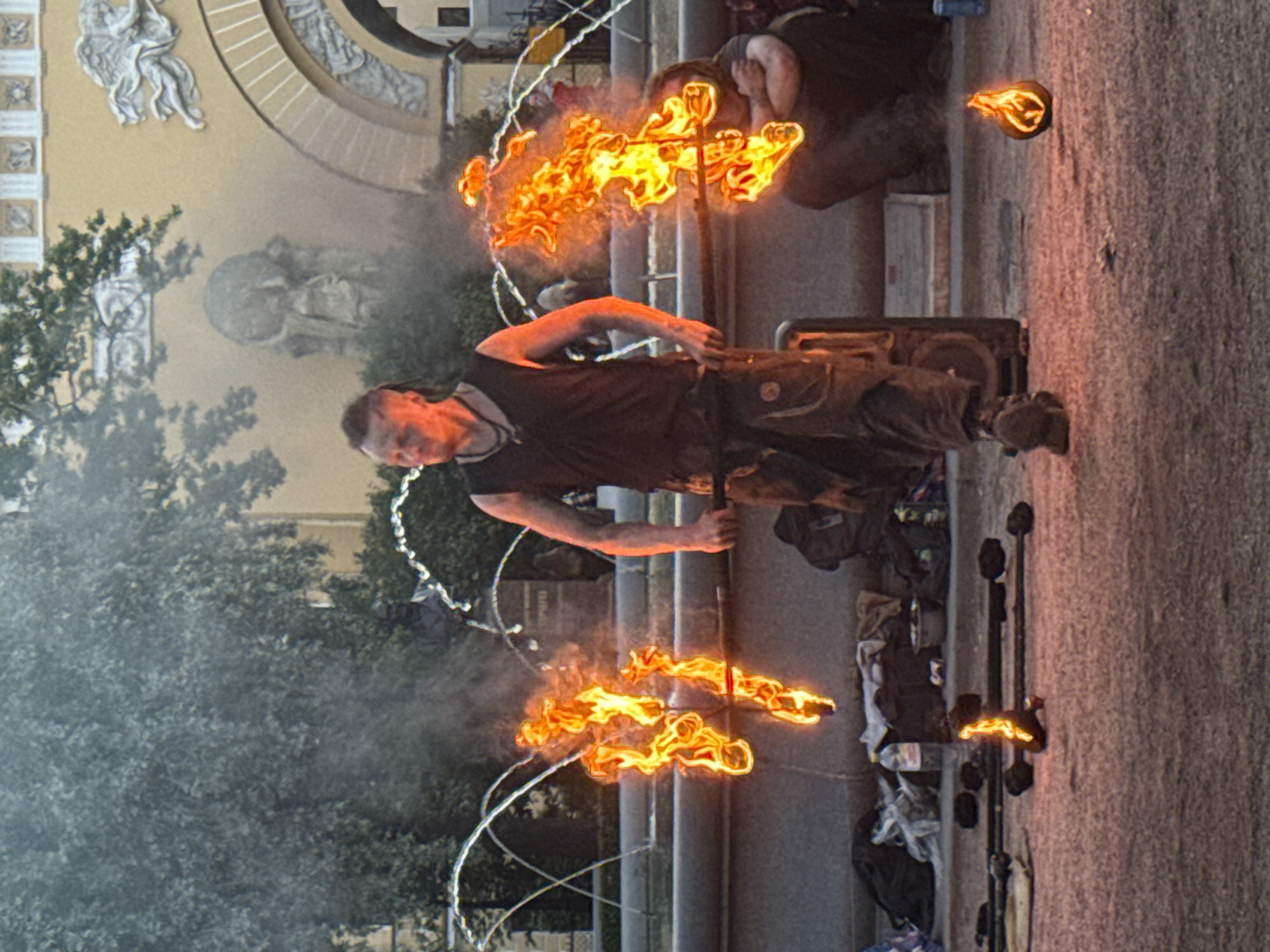 A male performer with dark hair styled in a bun is twirling burning poi in front of a fountain in Saint Petersburg, Russia. He is wearing a dark tank top and brown pants. Another person, whose gender is not definitively identifiable, is seated to his right, also with their arms crossed. The background features a building with classical architecture, adorned with sculptures and arches, and a treed area. The fountain is spraying water in arcs behind the performer. The ground is paved with a textured surface, and various items, including bags and a bottle of water, are scattered around the area. A small fireball is visible on the ground in the foreground, to the right. The scene suggests an outdoor performance or demonstration.