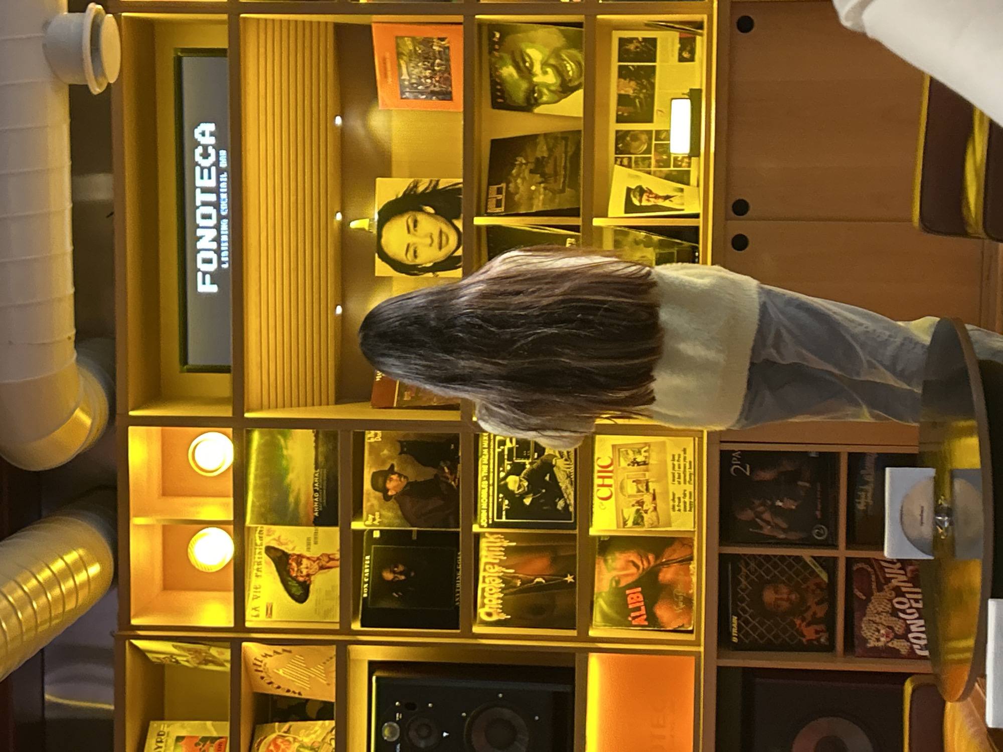 An indoor scene in Yerevan, Armenia, depicts a person with long, dark hair, wearing a light-colored top and blue jeans, standing with their back to the camera, seemingly browsing an extensive collection of vinyl records or album covers. The setting is a "FONOTECA LISTENING COCKTAIL BAR," as clearly indicated by a vertically oriented, illuminated sign on the left side of the custom-built wooden shelving unit.
The shelves are meticulously organized into individual compartments, each showcasing album artwork. Warm, yellow-orange spotlights are embedded within the shelves, illuminating the records and creating an inviting, ambient glow. Notable album titles and artist names visible include "AHMAD JAMAL," "LA VIE PARISIENNE," "BON CARTER," "Chocolate Milk," "JOHN MORALES THE MEM MIX," "ALIBI," "CHIC," "2PA" (likely 2PAC), "B TRAIN," and "CONGO LINK." A large speaker is placed on a lower shelf to the left.
On the far left, industrial-style ventilation pipes, some white and others golden-bronze, run vertically, contrasting with the rich wood tones of the shelving. The artificial lighting throughout the space suggests an evening or nighttime setting, typical for a cocktail bar environment designed for music appreciation. The overall atmosphere appears relaxed and curated for an immersive listening experience.