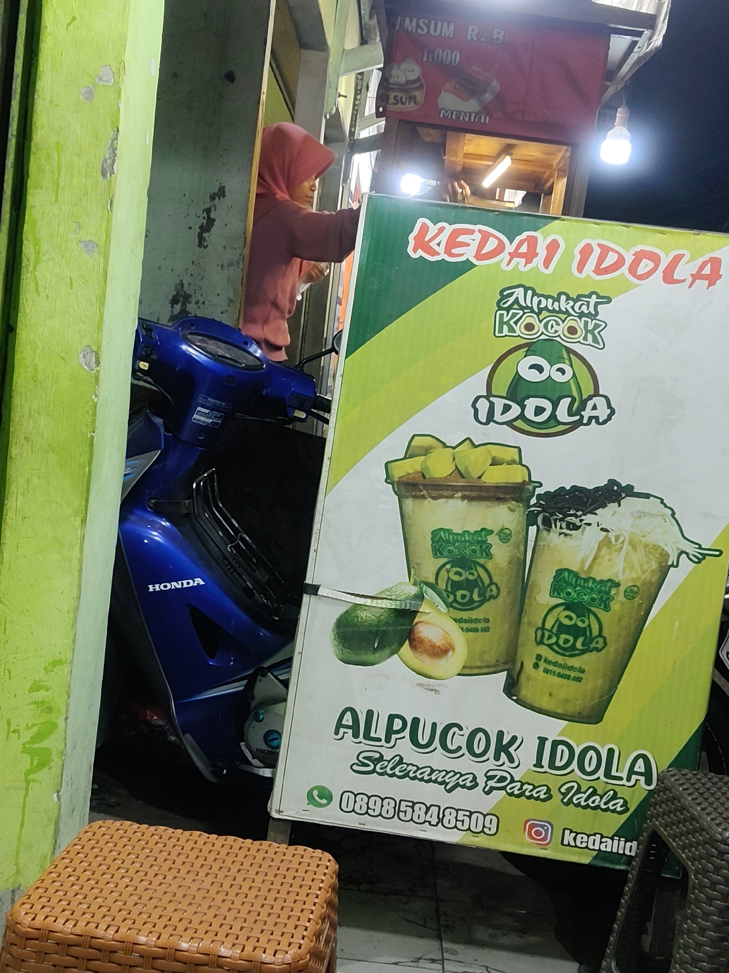 The image shows a street-side food stall in Mekarmukti, Indonesia, specializing in avocado drinks. A blue Honda motorcycle is parked in front of the stall, partially obscured by a green wall. A person wearing a pink hijab stands behind the stall, preparing drinks. A large banner advertises "ALPUCOK IDOLA," featuring images of avocado smoothies and contact information. The stall appears to be open for business, with a bright light illuminating the workspace. The overall scene depicts a typical evening setting at a local food vendor in Indonesia.