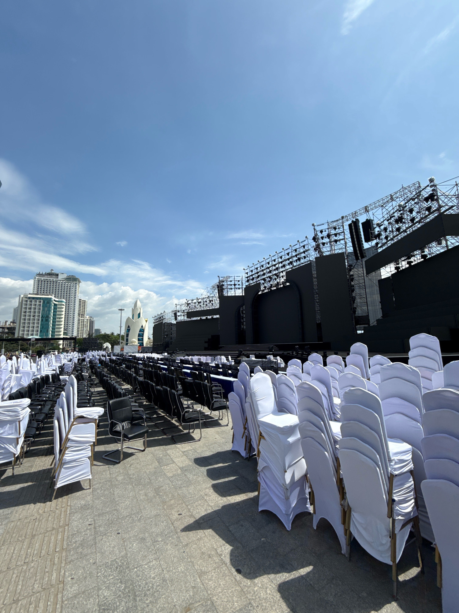 The image depicts an outdoor event setup in Nha Trang, Vietnam, under a clear blue sky with scattered clouds. In the foreground, rows of empty chairs are neatly arranged. Some chairs are covered with white fabric, creating a formal appearance, while others are black. Behind the chairs, a large, dark stage structure dominates the mid-ground. This stage is equipped with multiple large screens and a complex lighting and sound system, suggesting it is set up for a concert, performance, or public address. In the background, modern high-rise buildings are visible, indicating the location is within an urban area of Nha Trang. A distinctive white building with a pointed, sail-like roof is also present, adding architectural interest to the cityscape. The overall scene is one of preparation for a large-scale event.