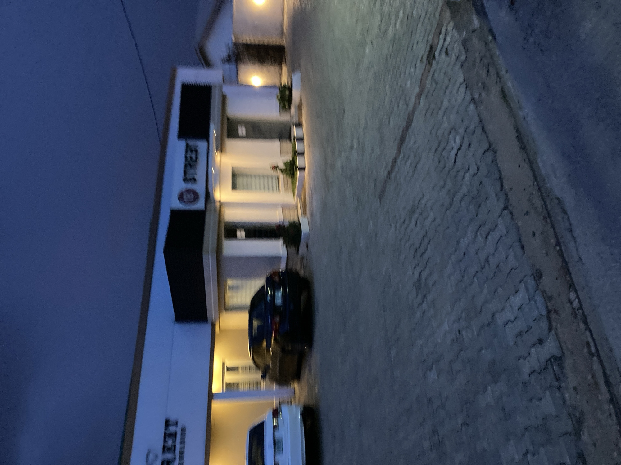 The image depicts a building facade at dusk, likely in Jos, Nigeria. The building appears to be a commercial or residential complex, with a modern design featuring white walls and dark accents. A prominent sign on the building reads "STREET".
Two cars are parked in front of the building: a dark blue sedan and a white van. The blue sedan is positioned closer to the building, while the white van is further away. The pavement in front of the building is paved with interlocking bricks.
The sky is a deep blue, indicating that the sun has set, and artificial lights from the building and street lamps are illuminating the scene. The overall atmosphere is calm and quiet, suggesting a peaceful evening. There are no people visible in the image, and the setting appears to be a well-maintained urban or suburban area.
Notable details include the "STREET" signage, the types of vehicles, and the paved ground. The time of day is clearly evening or dusk, and the weather appears to be clear. The location cues are limited to the possibility of it being Jos, Nigeria.