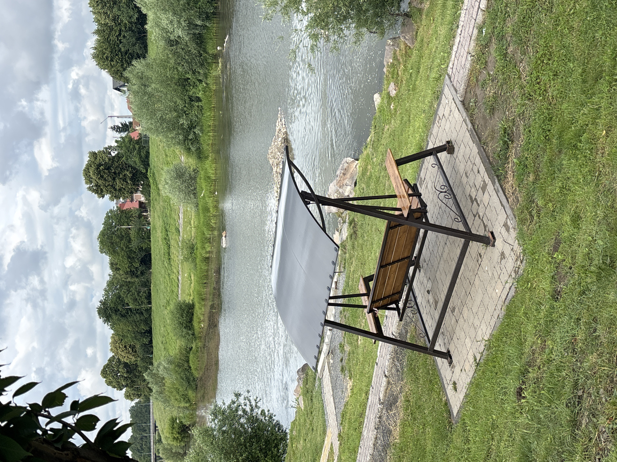 An outdoor scene on a cloudy day features a metal swing set with a wooden seat and backrest situated on a paved patio. The patio is bordered by green grass and a stone pathway leading towards a body of water. The riverbank is lined with green foliage, and across the water, a grassy hill is dotted with trees and houses, suggesting a semi-urban or suburban environment. A bridge is visible in the distance. The scene appears tranquil and serene, with no people present.