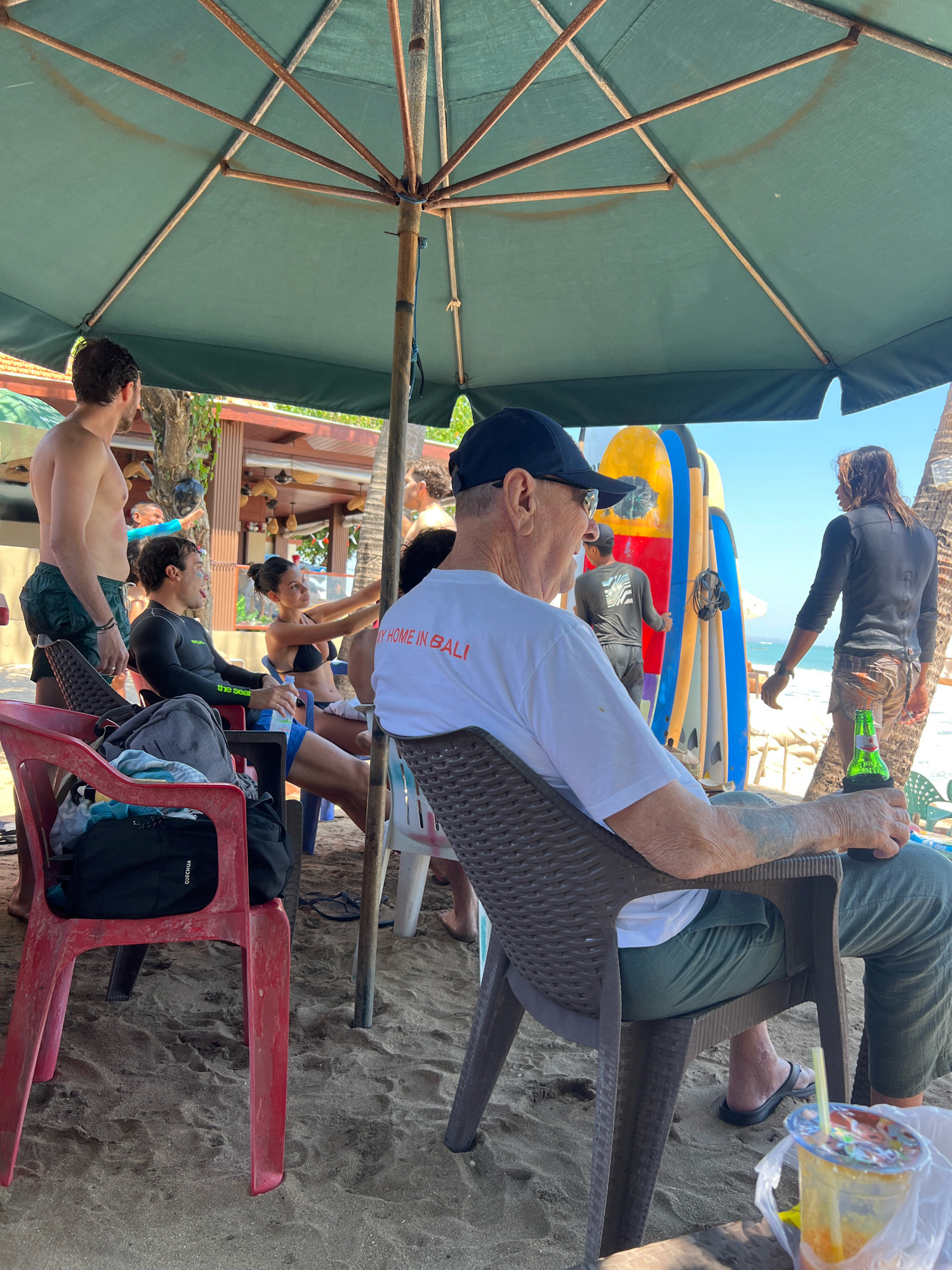 A group of people are gathered on a sandy beach under a large green umbrella. In the foreground, an elderly man wearing a white t-shirt with "MY HOME IN BALI" printed on it and green pants sits in a dark brown woven plastic chair, holding a green bottle of beer. Next to him on a low table is a plastic cup with a straw containing an orange drink. To his left, a red plastic chair is occupied by a person with a backpack on it. To the left of the red chair, a man in green shorts and a dark rash guard sits in a blue chair, with a bottle of water in his hand. Behind him, a man with dark curly hair and green shorts stands. Further back, a woman in a black bikini is talking to a man. In the background, several surfboards are leaning against a structure, and a person in a dark shirt and shorts walks away from the camera. The ocean is visible in the distance. The scene appears to be in Kuta, Indonesia, given the surfboards and beach setting.