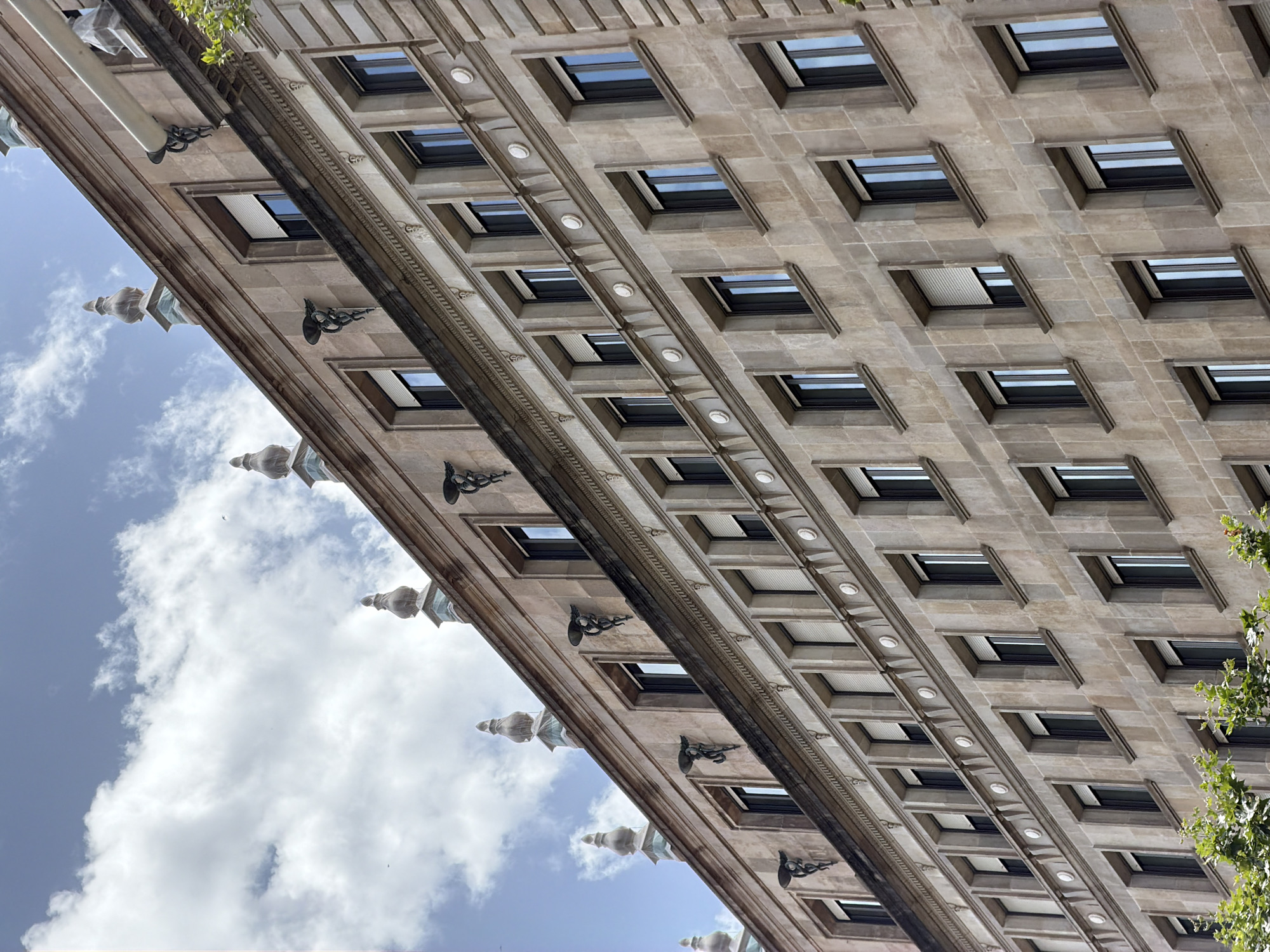 The image shows the facade of a building in Barcelona, Spain. The building is made of light-colored stone and features a repetitive pattern of rectangular windows. The windows are dark, suggesting they are either covered or tinted. The architectural style appears to be classical or neoclassical, with decorative elements such as moldings and what look like small statues or gargoyles between some of the windows.
The perspective is from below, looking upwards at an angle, which emphasizes the height and scale of the building. The sky is visible in the upper left portion of the image, filled with white, fluffy clouds against a blue background. This suggests it is daytime, likely midday or afternoon, with fair weather. A few birds are also visible in the sky.
The image is a still shot, with no discernable activity or event taking place. The focus is entirely on the building's architecture and its relationship with the sky. There is no visible text in the image. The overall mood is one of architectural grandeur and the serenity of a clear day.