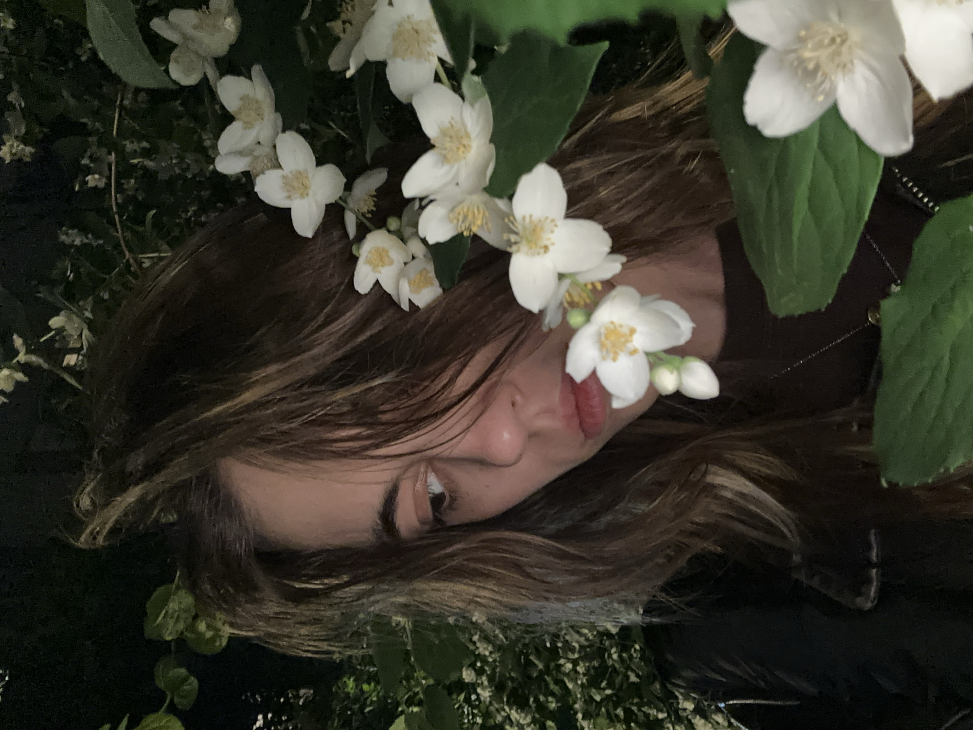 In Moscow, Russia, a young woman with long, wavy brown hair and light brown eyes is captured in a close-up, self-taken photograph. She is positioned amongst lush green foliage and delicate white jasmine flowers, some of which are in the foreground, partially obscuring her face and body. Her head is tilted downwards and to her right, with her gaze directed away from the camera. She wears a dark, possibly black, jacket over a dark-colored shirt, with a subtle necklace visible. The background consists of dark, out-of-focus greenery and hints of more white flowers, suggesting an outdoor setting at night or in low light conditions. The overall ambiance is intimate and serene.