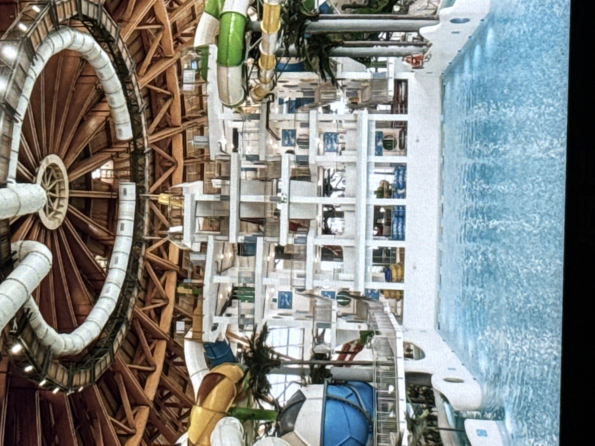 The image shows an indoor water park with multiple levels of slides, pools, and walkways. The structure has a large, domed wooden roof with exposed beams. Water slides in yellow, blue, and green are visible, along with a large blue pool in the foreground. The architecture appears to be modern, with white structures and metal railings. Palm trees are also present, suggesting an attempt to create a tropical atmosphere indoors. There are no people clearly visible in the image. The water park is likely located in the city of Norilsk, Russia, a city known for its harsh Arctic climate, making indoor recreational facilities like this essential.
