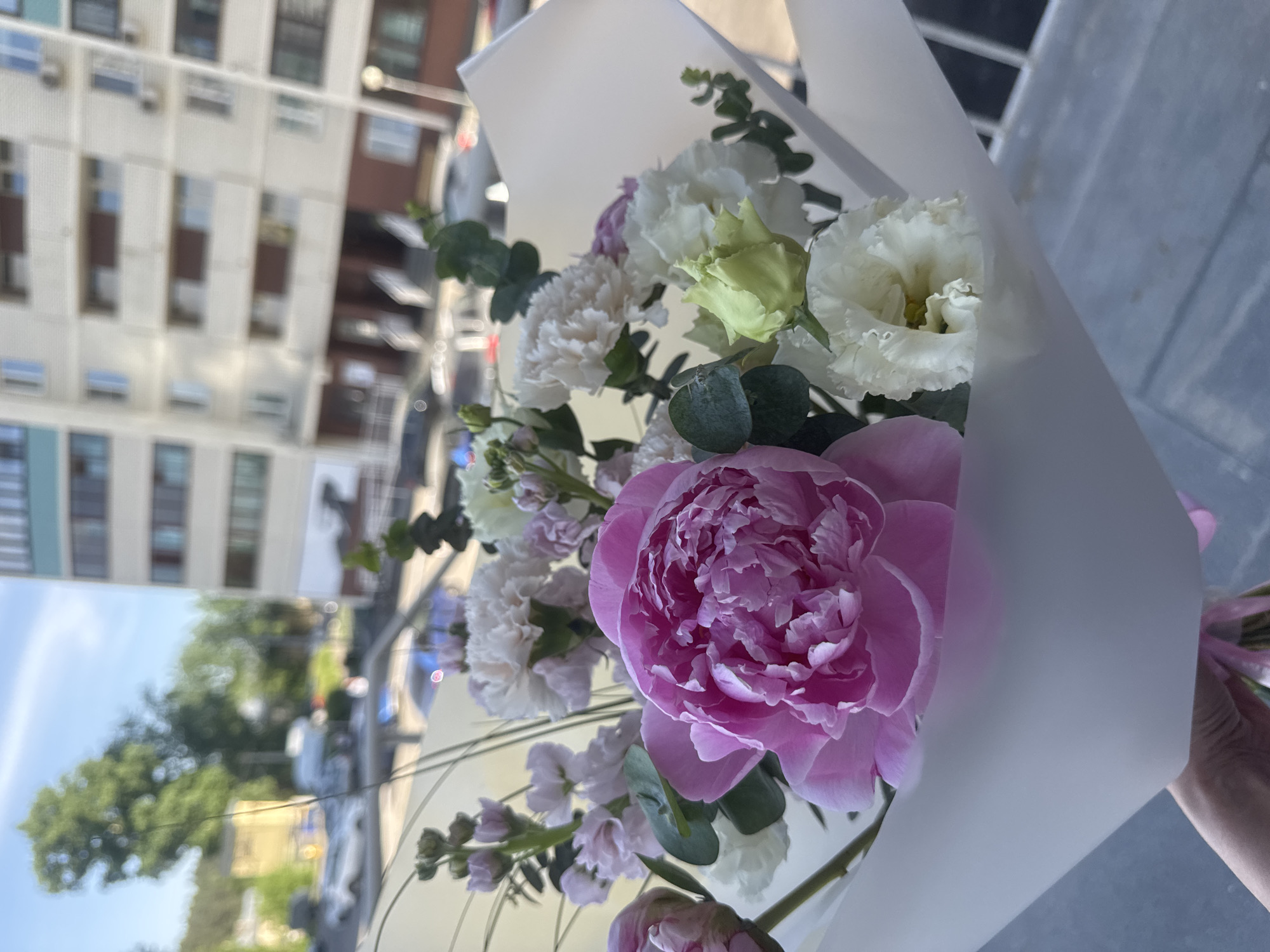 A bouquet of flowers is held in front of a city scene. The bouquet prominently features a large pink peony and other white and light-colored flowers, wrapped in white paper. The background shows a city with buildings, cars, and trees, suggesting an urban environment. The location is Izhevsk, Russia.