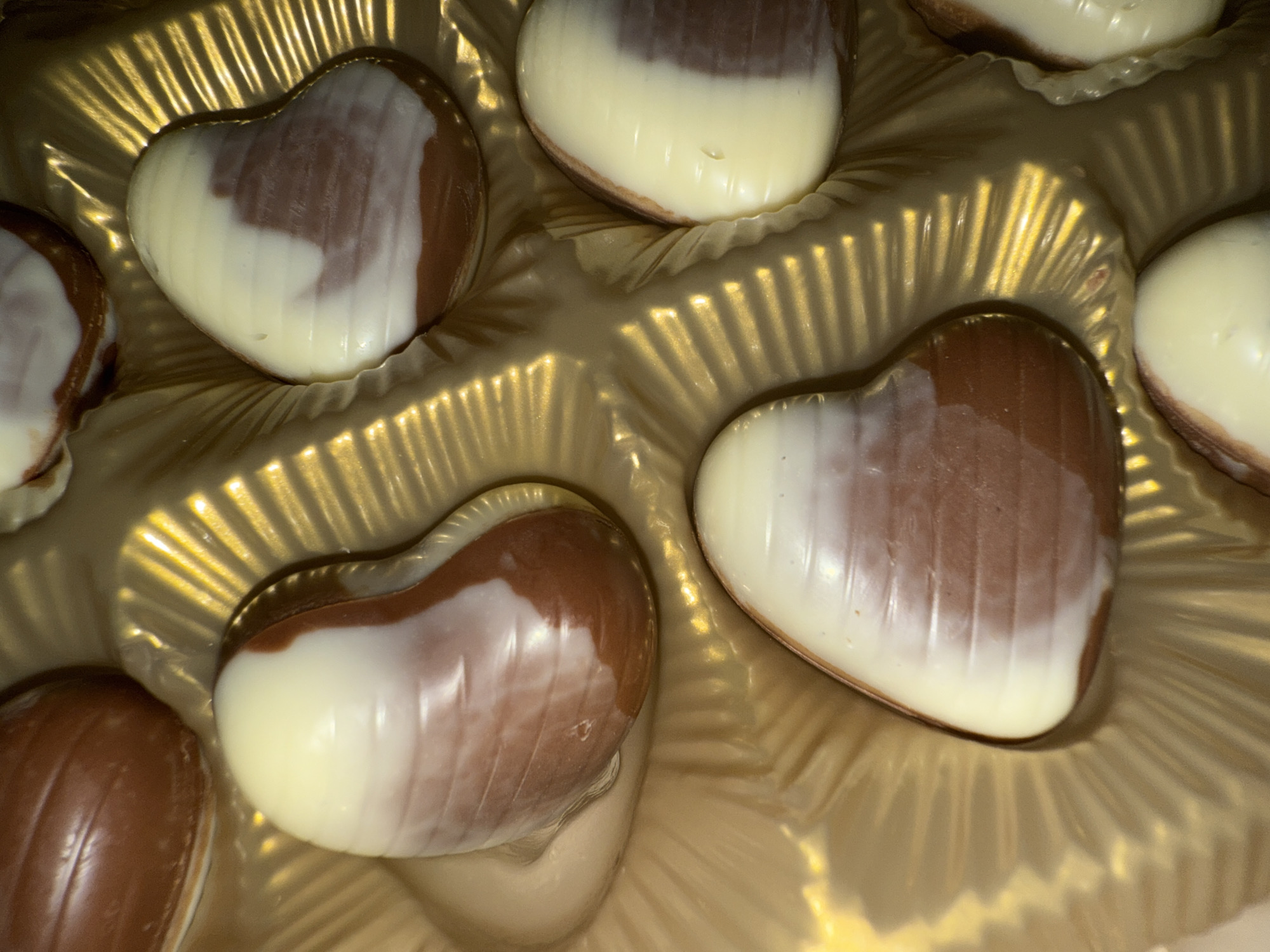 The image displays an overhead view of several heart-shaped chocolates arranged in a golden, compartmentalized box. Each chocolate is a blend of dark and white chocolate, with the white chocolate appearing as swirls or distinct layers over the rich brown of the dark chocolate, creating a marbled or two-toned effect on their smooth, glossy surfaces.
The chocolates are nestled within individual sections of the packaging, which is made of a shiny, reflective golden material, likely a molded plastic or foil insert. This material features a distinct ribbed or pleated texture, which generates prominent highlights and shadows under what appears to be warm, artificial lighting. This lighting enhances the golden sheen of the box and the inviting appearance of the chocolates.
Approximately six heart-shaped chocolates are visible in various stages of fullness within their compartments. No specific activity is taking place beyond their presentation in the box, suggesting they are a confectionery product, possibly a gift. No visible text is present on the chocolates or the packaging, and there are no discernible location cues within the image itself to indicate the context of Varna, Bulgaria. The heart shape inherently conveys themes of love or affection.