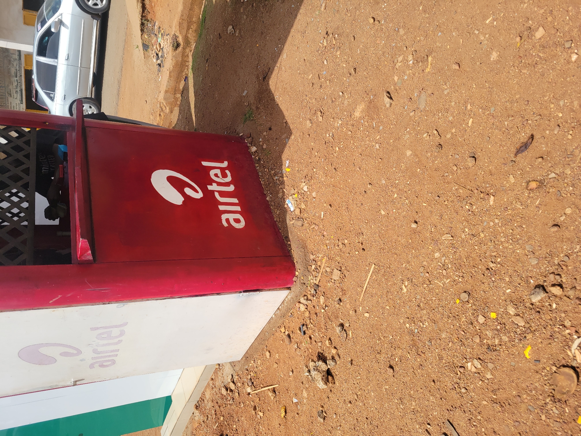 The image depicts an Airtel kiosk in Unknown, Unknown. The kiosk is predominantly red and white, with the "Airtel" logo prominently displayed. The surrounding environment is a dirt or gravel ground. A car is visible parked nearby. A person is also visible, partially obscured.