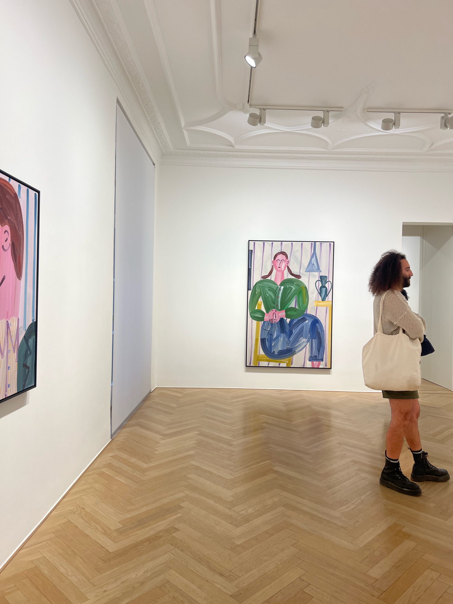 The image depicts the interior of a modern art gallery, likely in Berlin, Germany, based on the provided context. The room features white walls, a high, ornate ceiling with molding, and a polished wooden floor laid in a herringbone pattern. A person with curly dark hair, wearing a beige knitted sweater, olive green shorts, black socks, and black combat boots, stands facing right, observing a large, colorful painting on the wall. They are carrying a large, off-white canvas tote bag over their left shoulder. The painting features a stylized figure with exaggerated features, seated against a striped background. To the left of this painting, another framed artwork, partially visible, also shows a colorful, stylized portrait. Track lighting is installed on the ceiling, illuminating the artworks. The overall ambiance suggests a contemporary art exhibition space.
