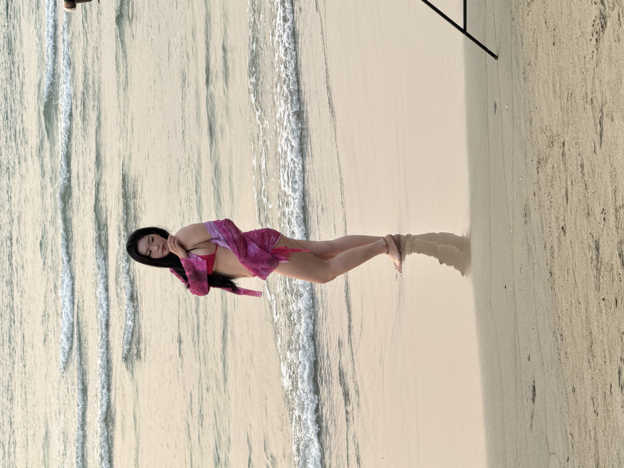 A woman lies on a sandy beach next to the ocean. She is wearing a pink bikini and matching sheer long-sleeved coverup. The beach has white sand and the water is a light blue. The location is likely Đà Nẵng, Vietnam.