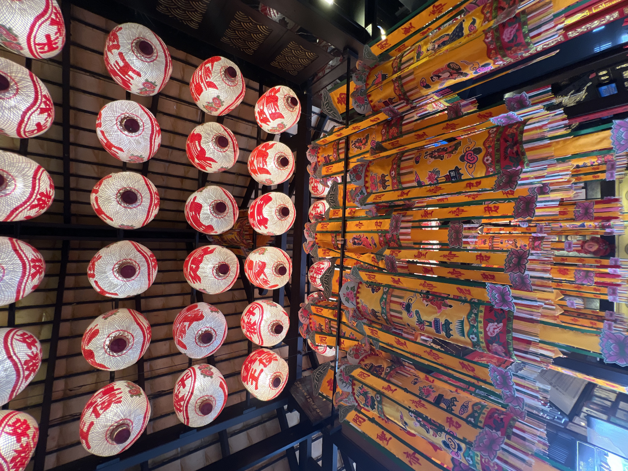 This is a low-angle, interior shot of a market stall in Baan Hua Thanon, Thailand, likely during a festival or special occasion. The ceiling is adorned with rows of white, oval-shaped lanterns with red decorative patterns. Below the lanterns, an abundance of long, yellow banners with red and green accents hang down. These banners are decorated with intricate imagery, including figures, calligraphy, and floral motifs. The scene suggests a vibrant and festive atmosphere, characteristic of local celebrations in Thai markets.